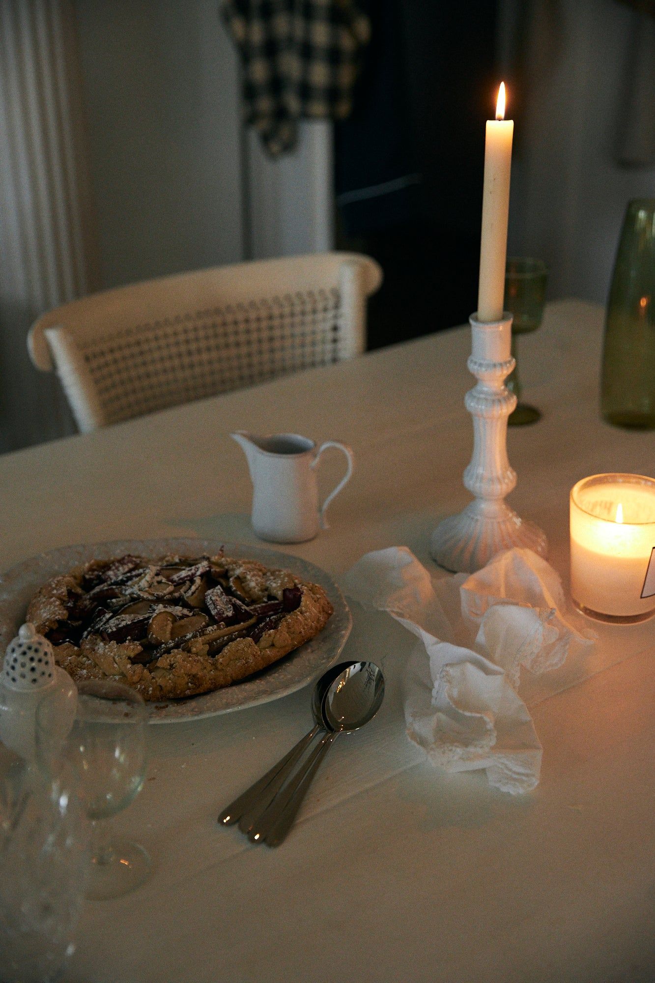 Table set with berry galette, lit candles, white creamer, napkins, silverware, empty glasses. Warm lighting.