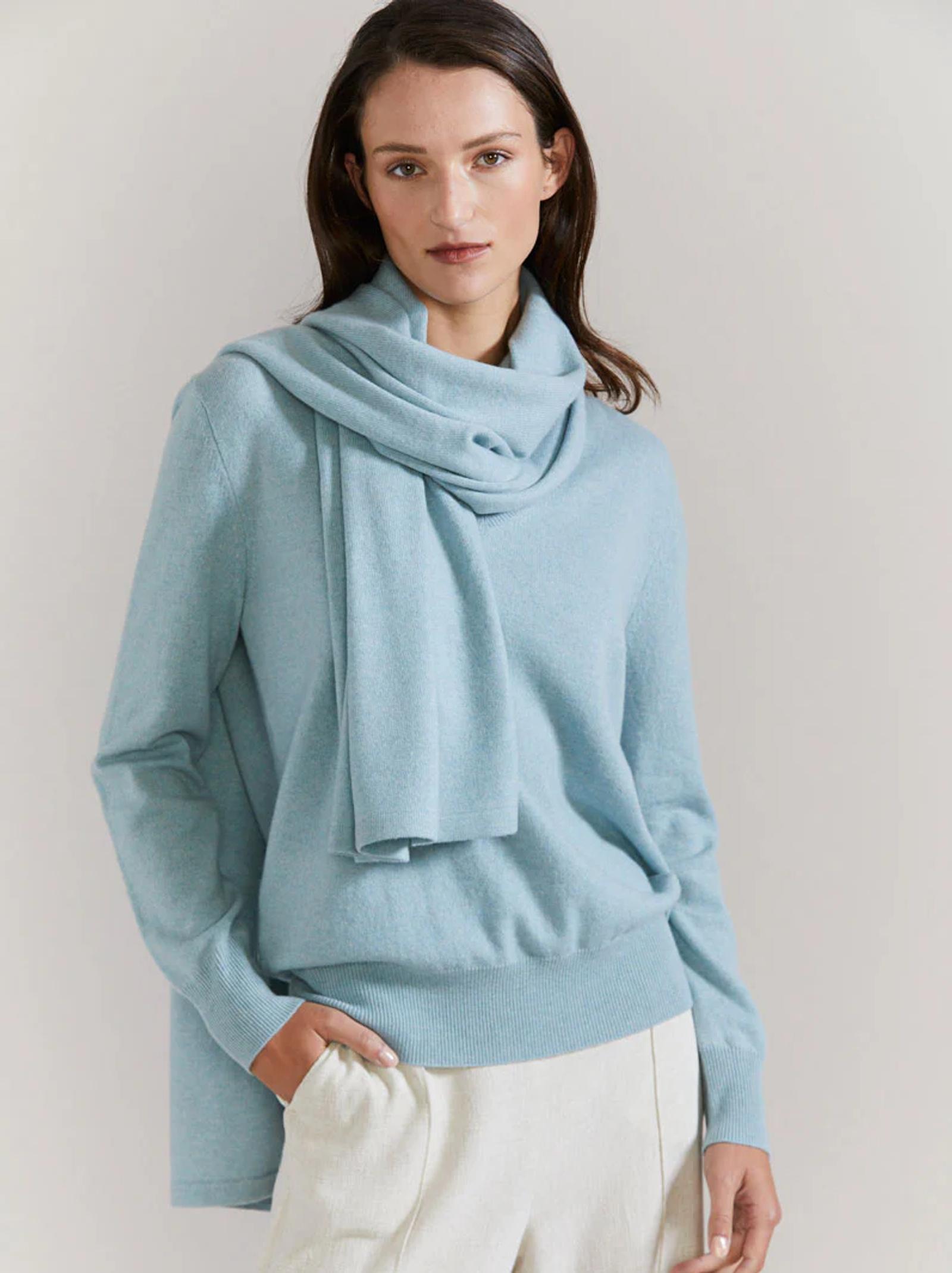 Dark-haired person in light blue sweater, scarf, and white pants. Standing against white background, hand in pocket, neutral expression.
