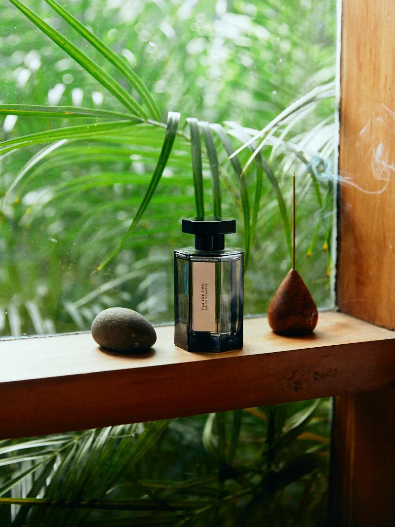 Glass perfume bottle with black cap on wooden window ledge. Two stones nearby. Green foliage outside.