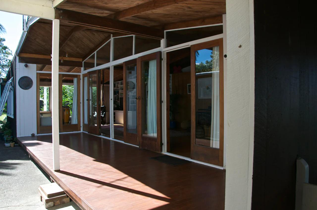 Wooden veranda with glass doors and white frames. Exposed beams over shaded seating area. Greenery and glimpse of home studio visible through doorways.