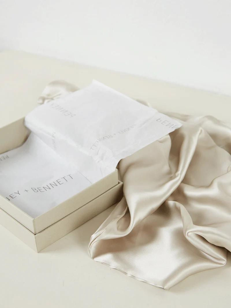 A beige silk garment out of an open cream-coloured gift box lined with tissue paper printed with the text "Penney + Bennett." Elegant and luxurious against a white background.