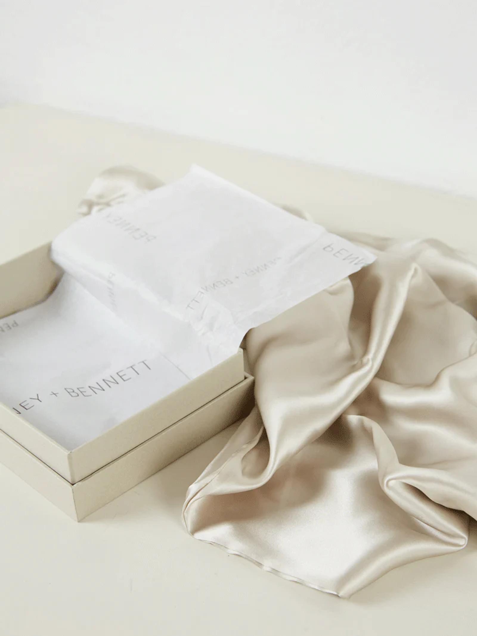 A beige silk garment out of an open cream-coloured gift box lined with tissue paper printed with the text "Penney + Bennett." Elegant and luxurious against a white background.