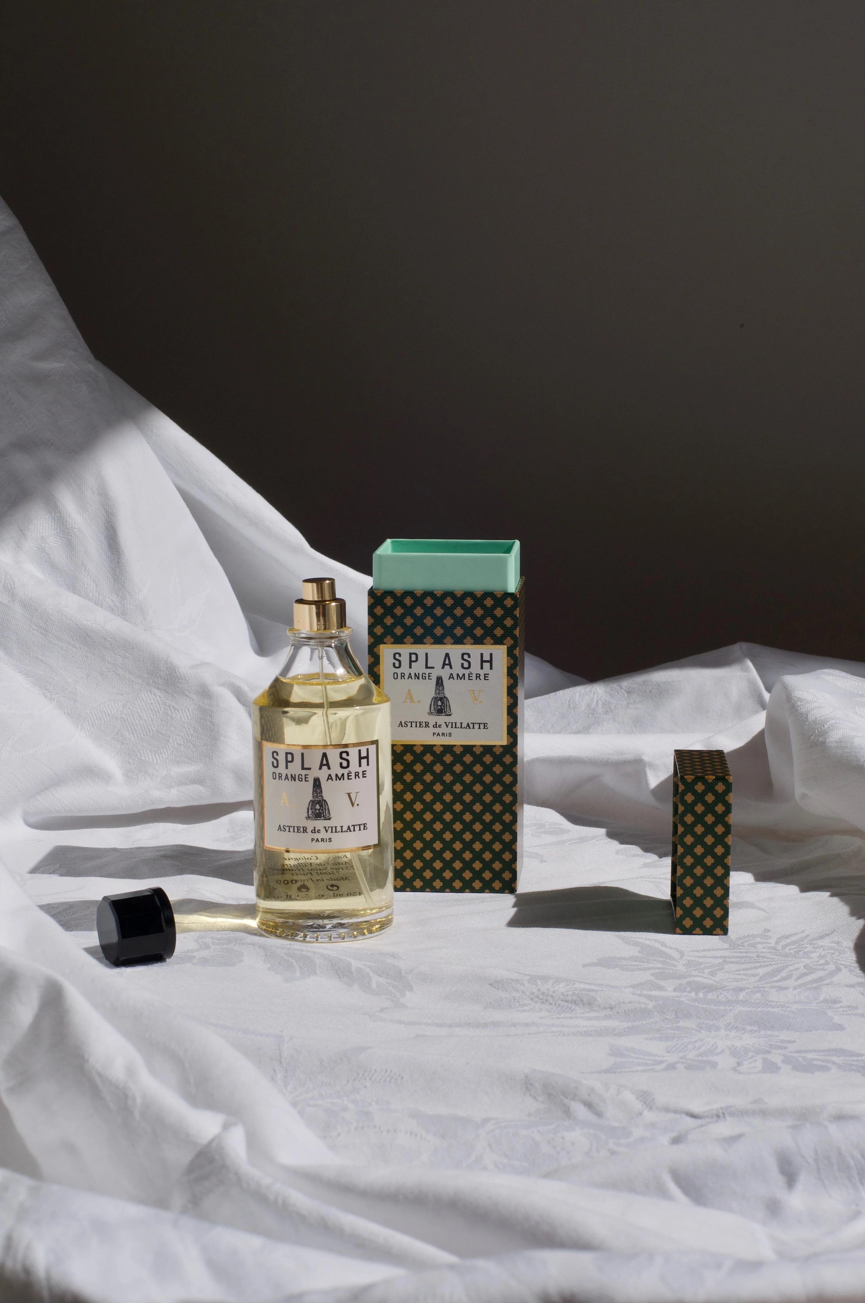 “Splash” Atelier de Villett parfum spray bottle with black cap beside it. Green and gold patterned square box is open behind the bottle on a white fabric surface.