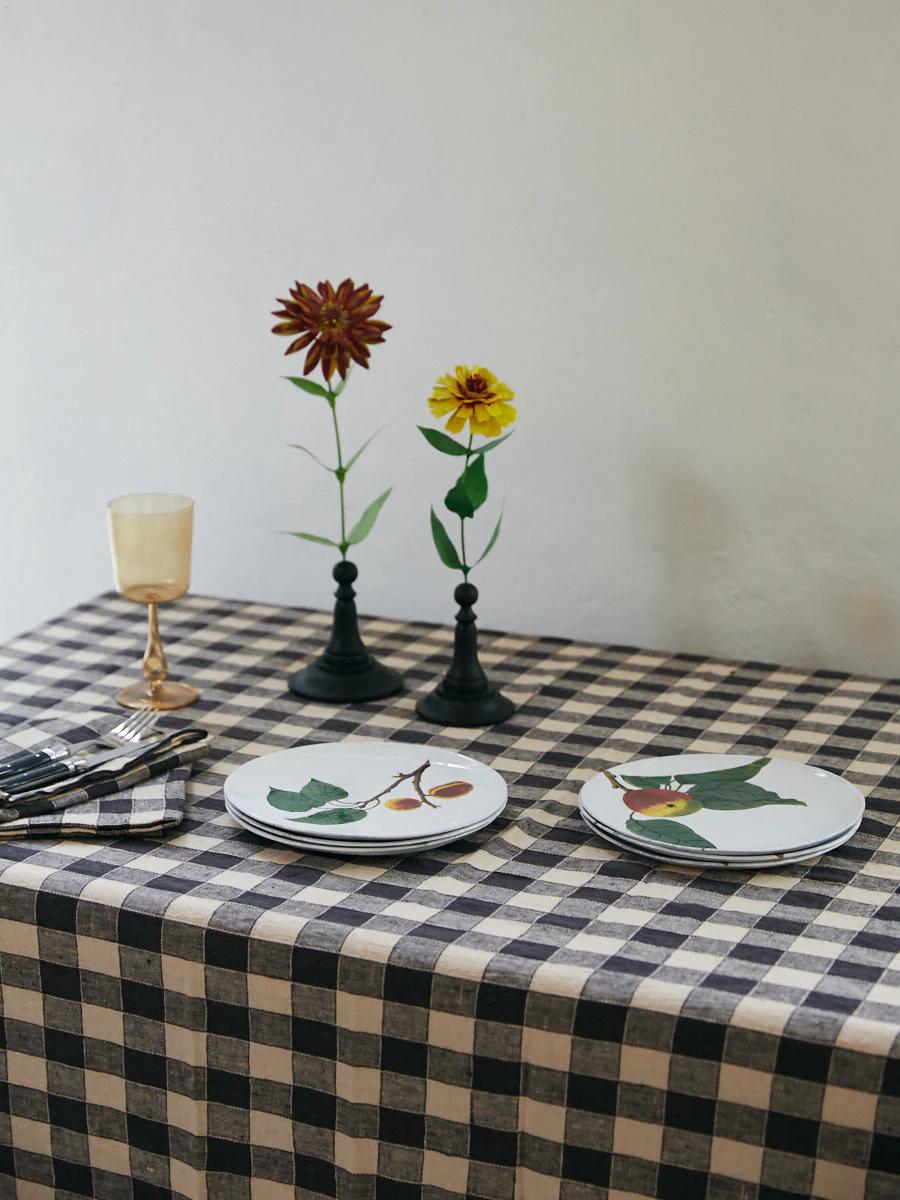 Dining table with black and white checkered linens. Botanical plates, silverware, beige glass, candle holders with flowers.