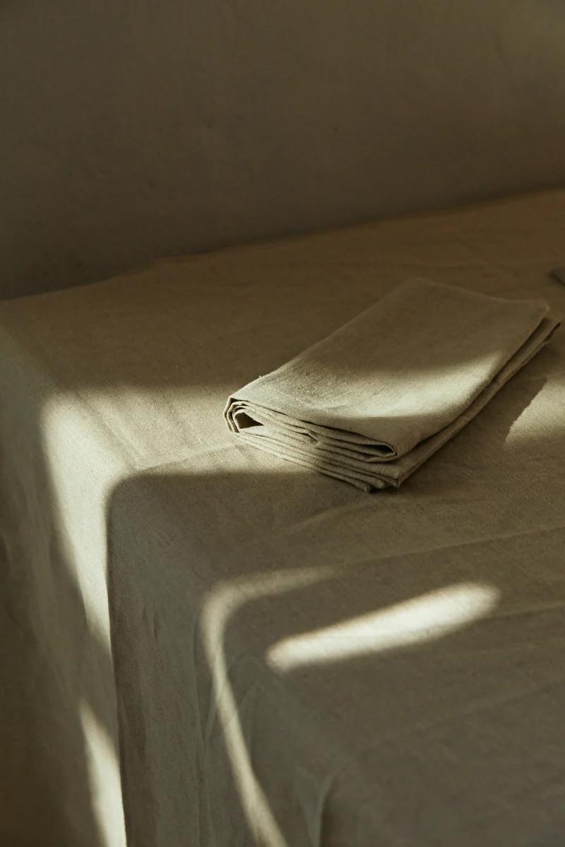 Beige Belgian Linen napkin on matching tablecloth. Sunlight creates warm shadows, serene atmosphere.