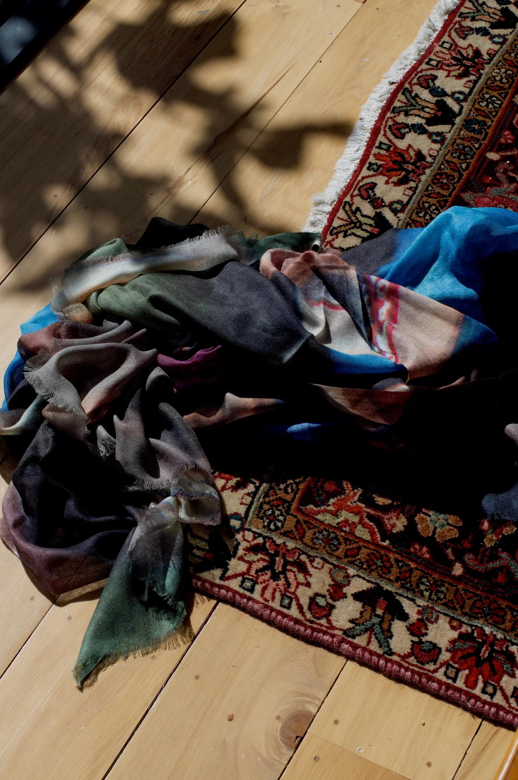 Colourful scarf on wooden floor next to fringed rug. Leaf shadows visible. Scarf features blue, pink, green, and grey hues.