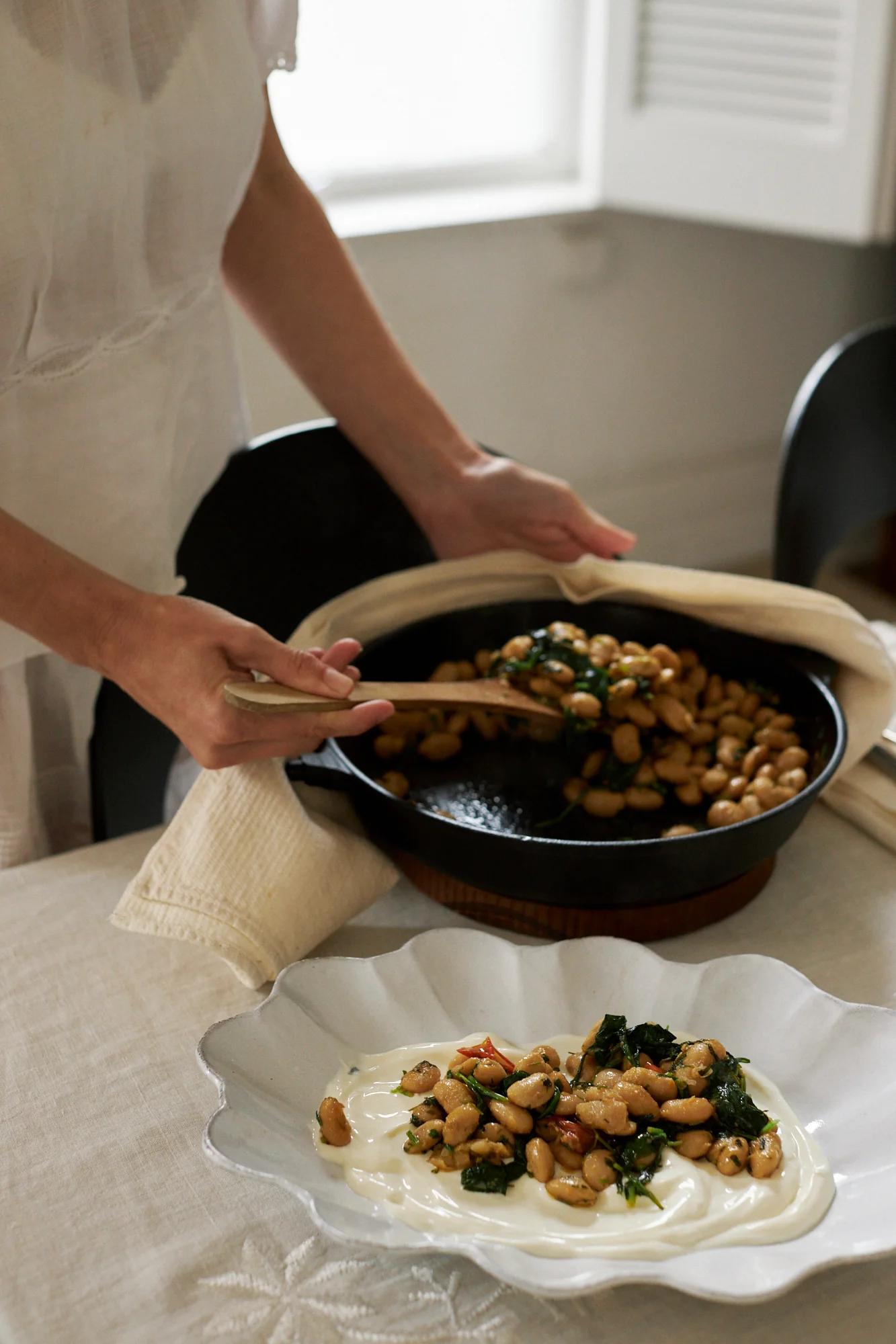 Someone in light clothing serves beans and greens from a skillet to a white plate. The table is set with a neutral tablecloth for Easter break.