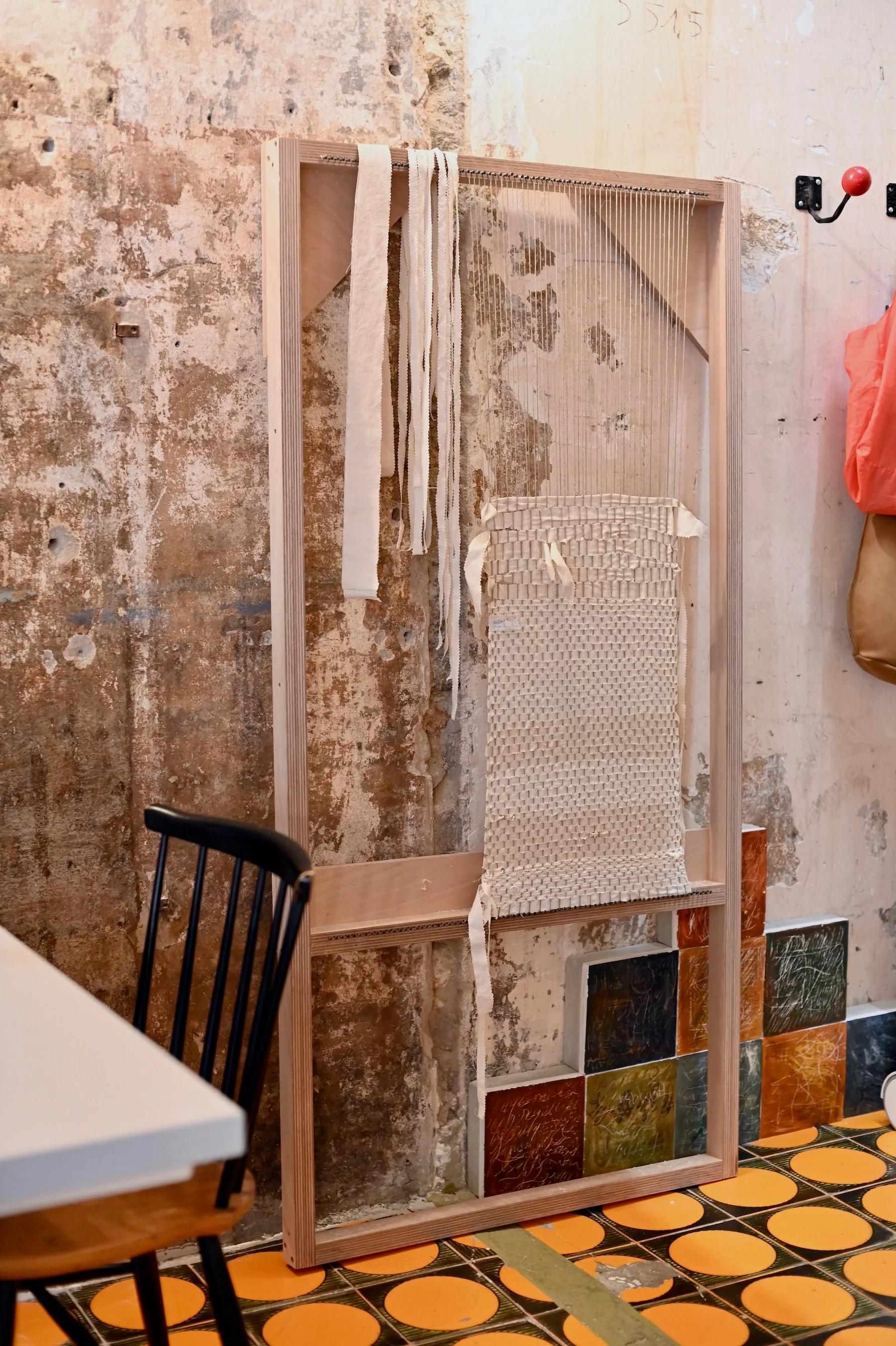 Unfinished weaving project on large wooden loom against distressed wall. Orange and yellow tiled floor, black chair nearby.