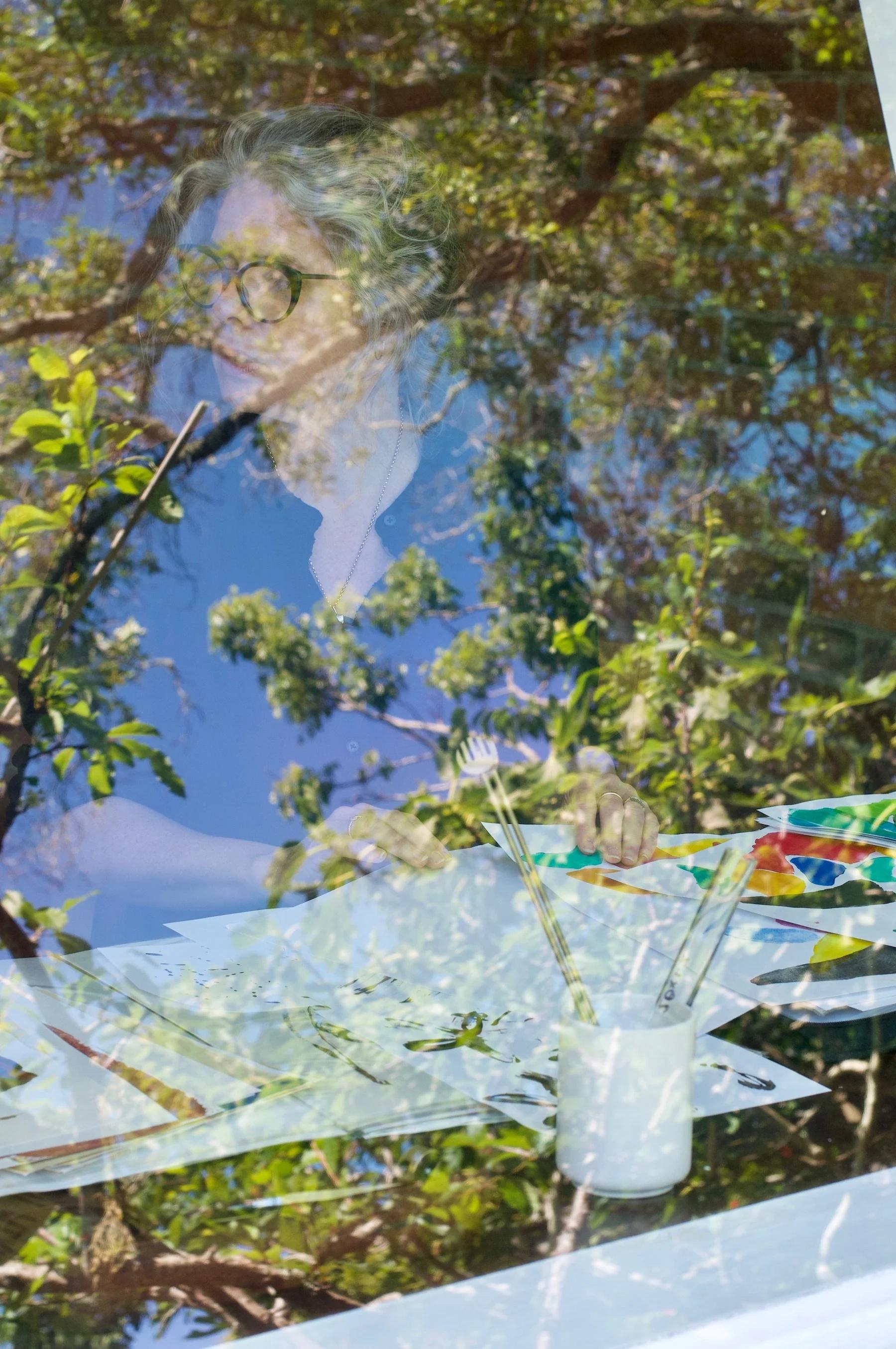 Person with glasses painting, seen through window. Tree and sky reflections overlay scene. Paintbrushes in container.