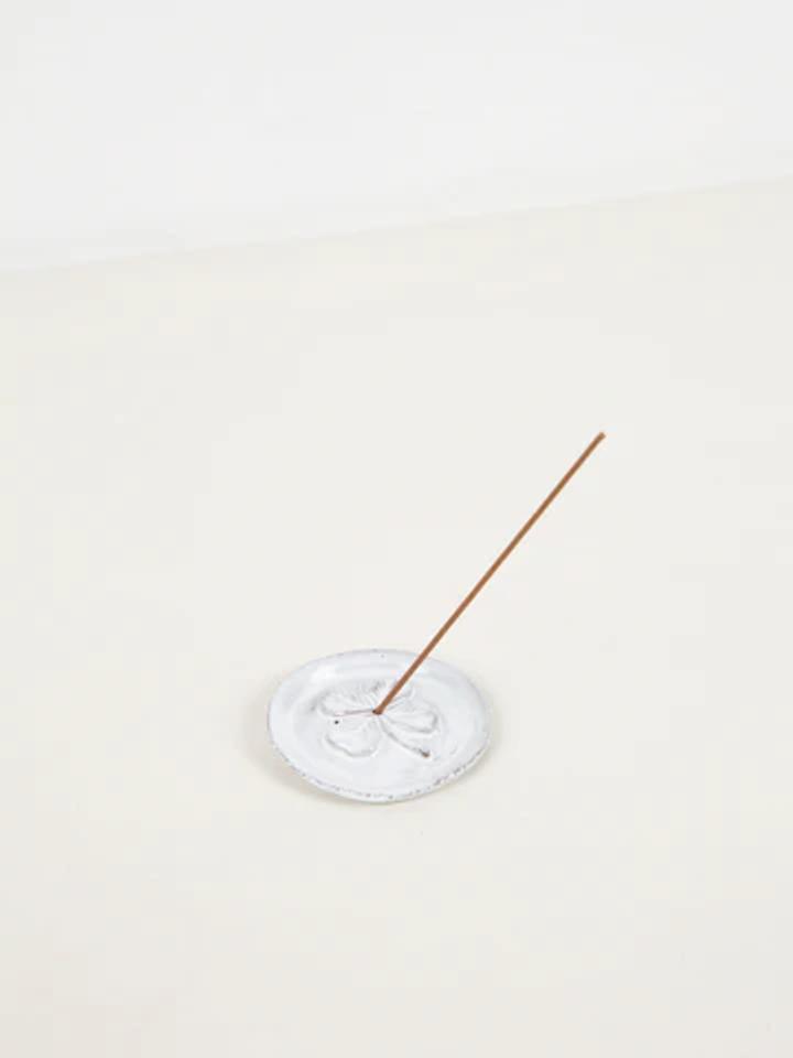 Thin Astier de Villatte incense stick in round holder emits wisp of smoke. Set against plain, light background, embodying understated elegance.