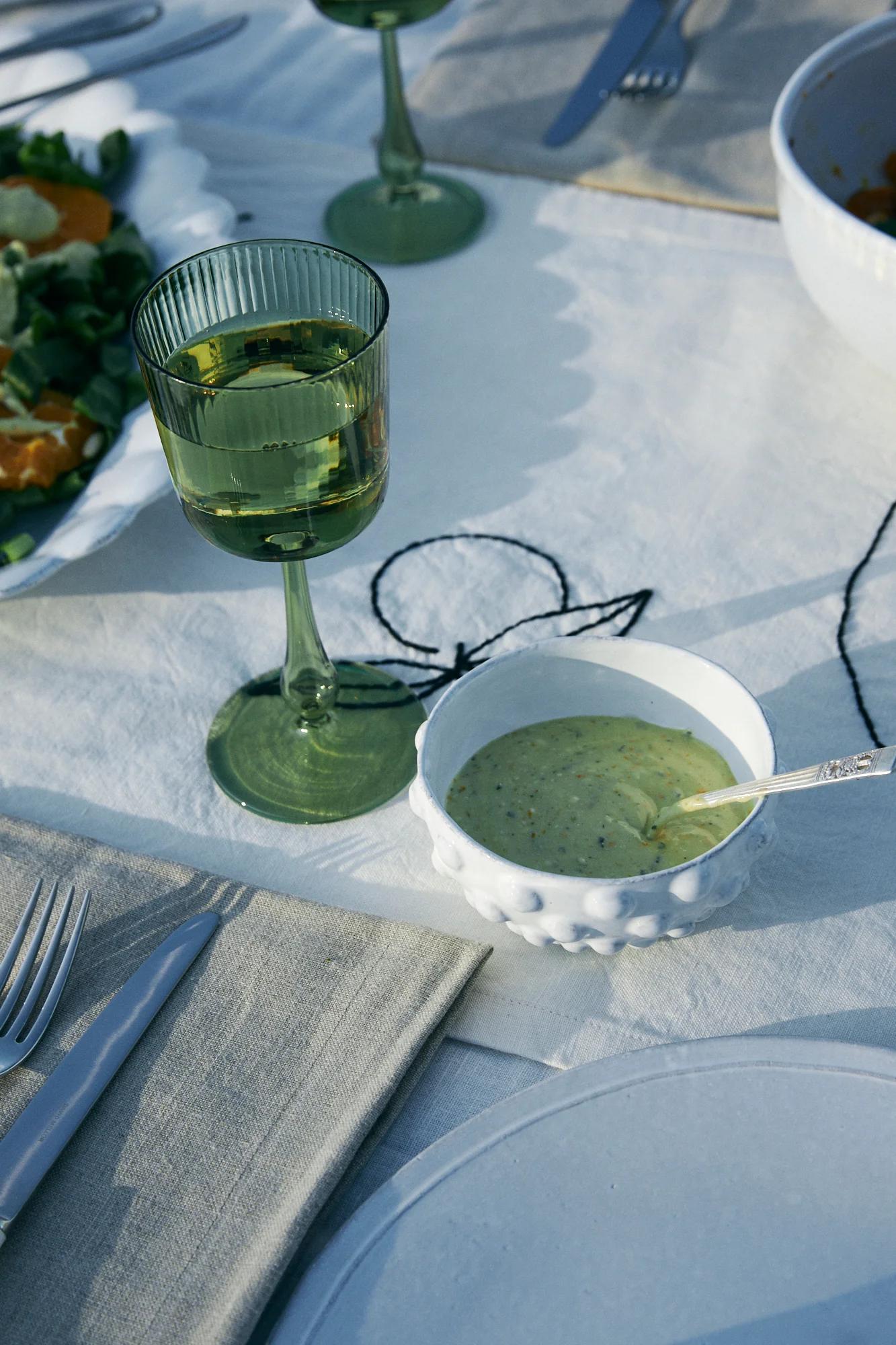 Table set with green wine glass, salad dressing bowl, plates and cutlery. Embroidered tablecloth.