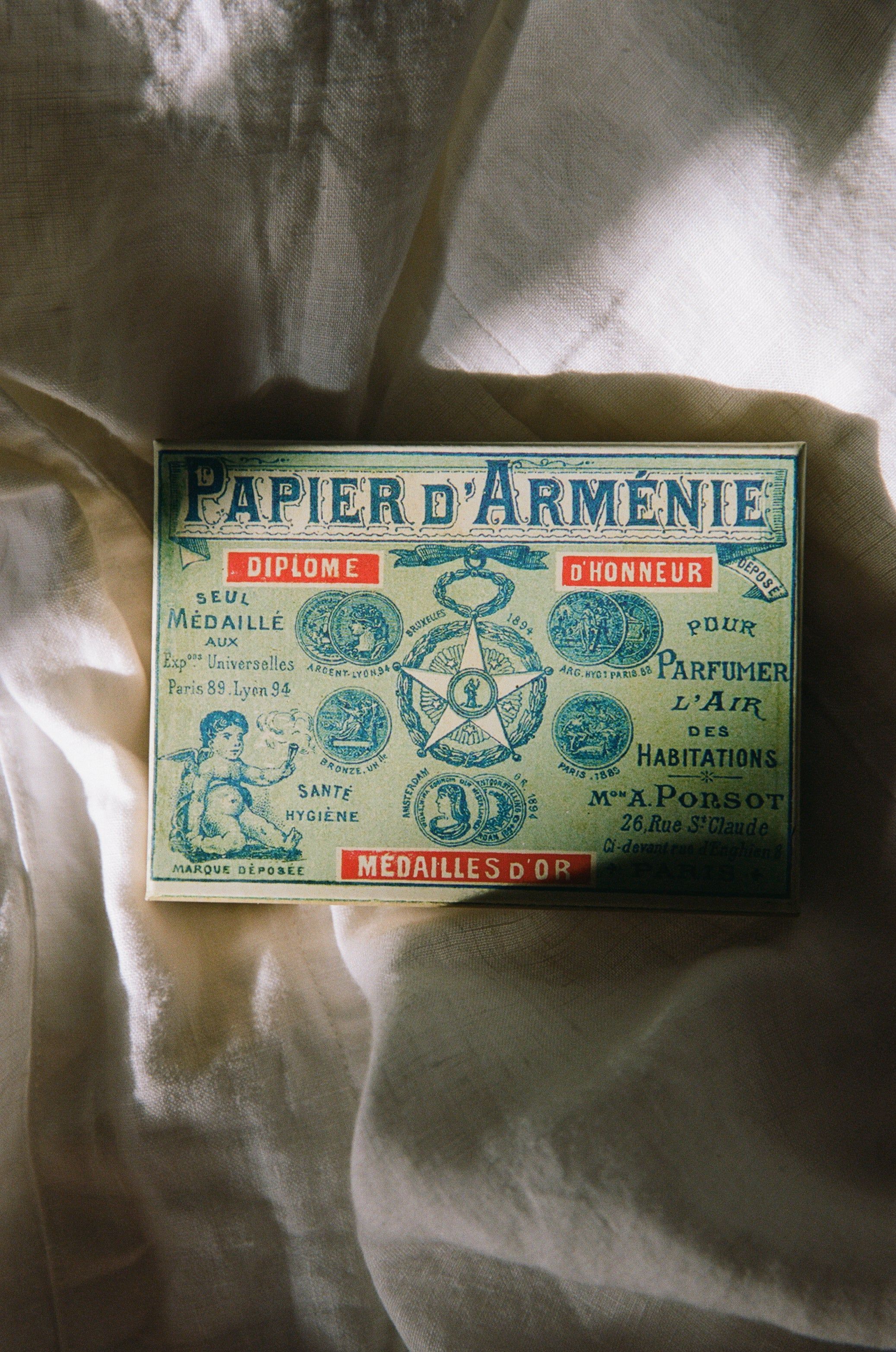 A vintage green Papier d’Arménie booklet rests on cream fabric. The intricate design features awards, medals, an anchor, and laurel wreaths, with French text by Auguste Ponsot.