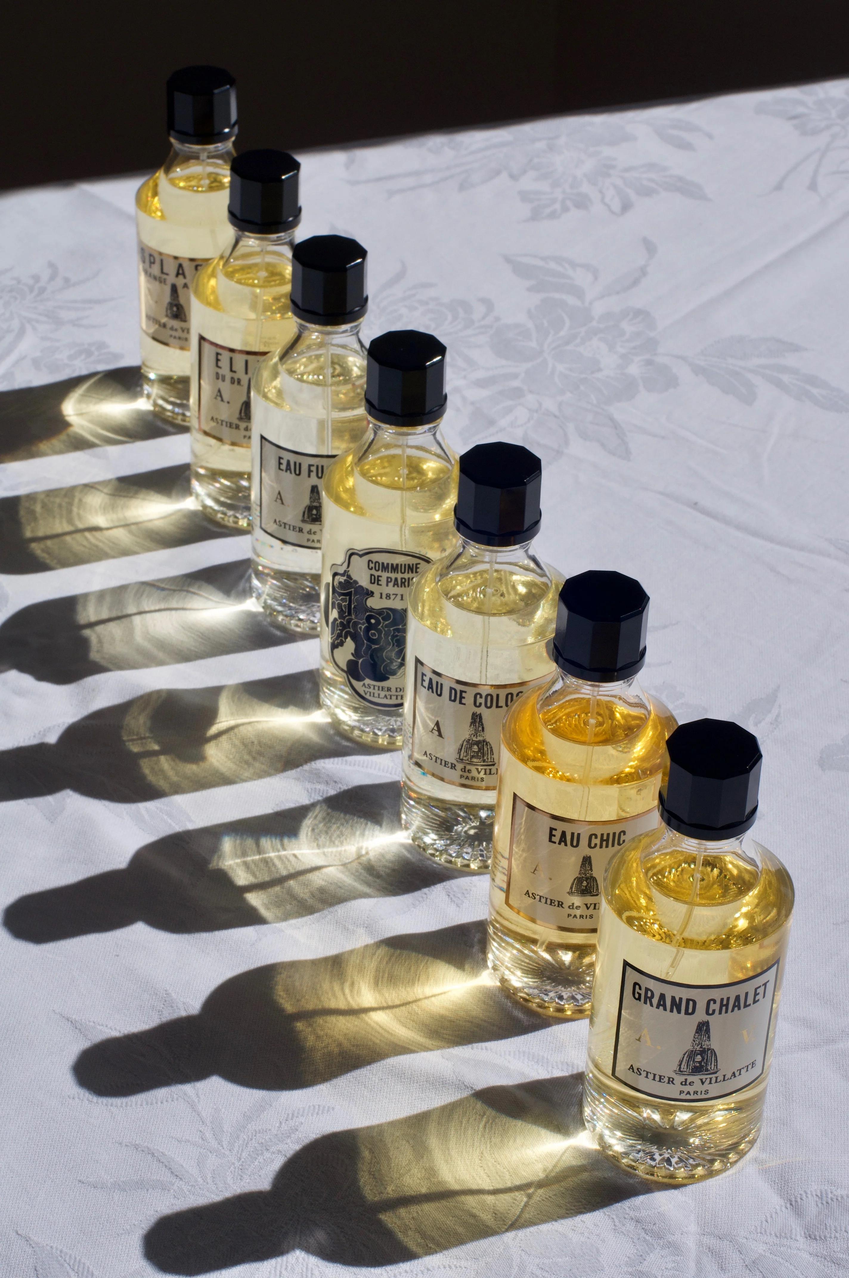 A row of seven Astier de Villatte colognes with distinct labels on a white tablecloth. Sunlight creates reflections under each bottle, showcasing their elegant, exclusive shades.