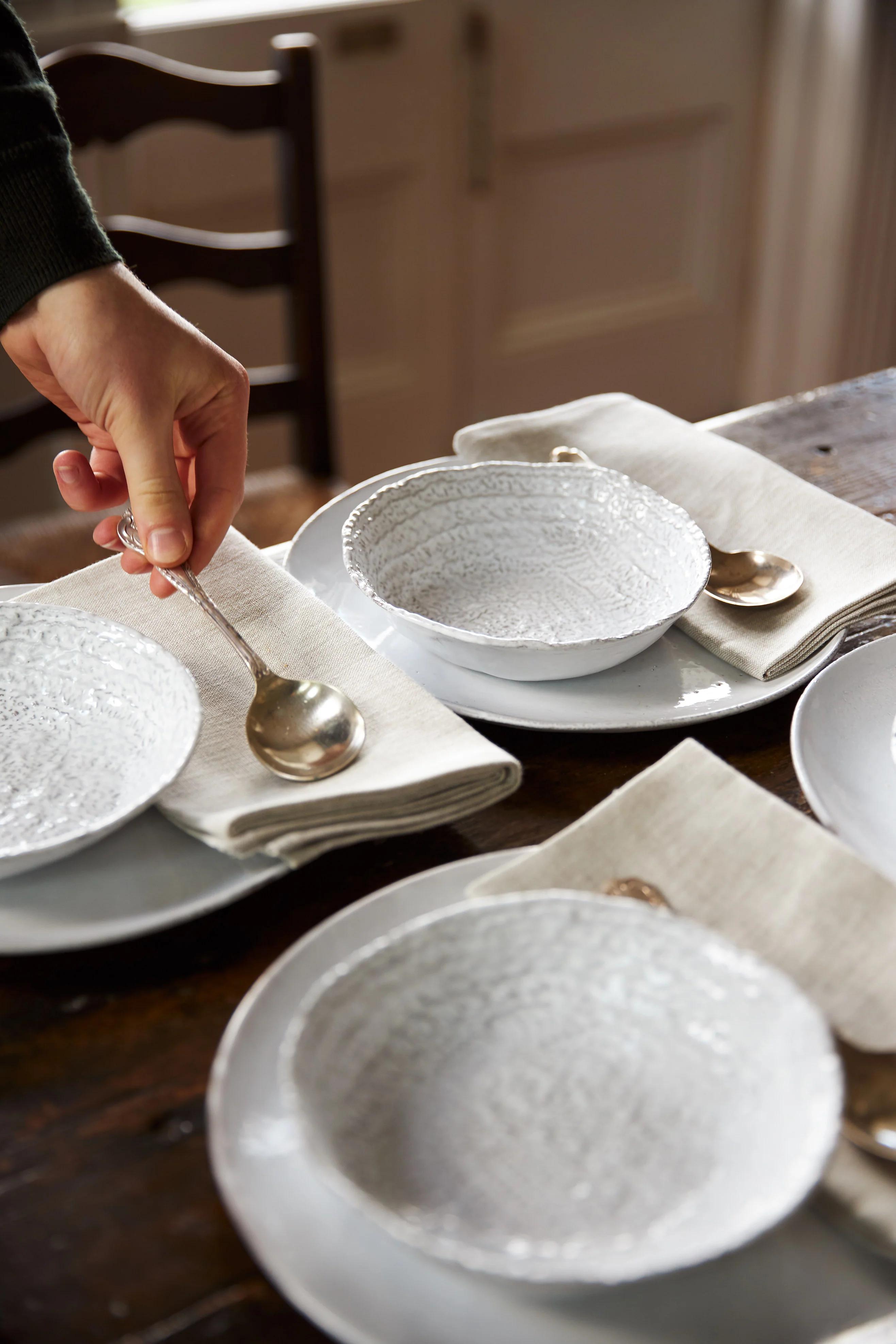 A hand places a silver spoon on a napkin next to an Astier de Villatte bowl. Several sets are prepared on a wooden table in natural light.