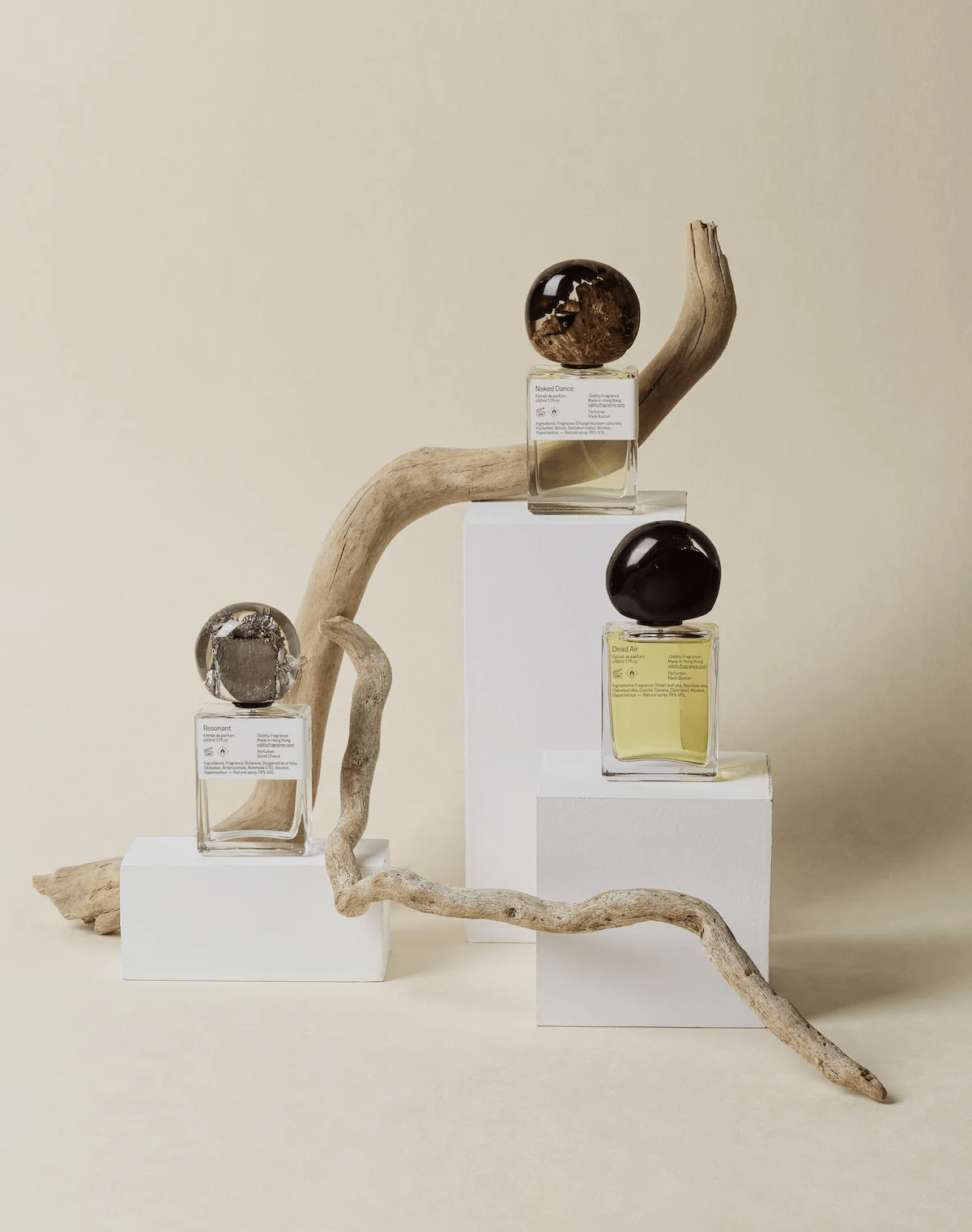 Three round-lidded perfume bottles on white platforms and driftwood. Labels visible, liquid colours vary from pale yellow to clear.