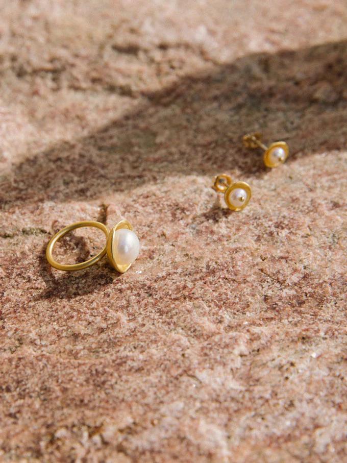 Gold pearl ring and pearl stud earrings on textured stone in natural light. Simple designs showcase jeweller's skill with organic materials.