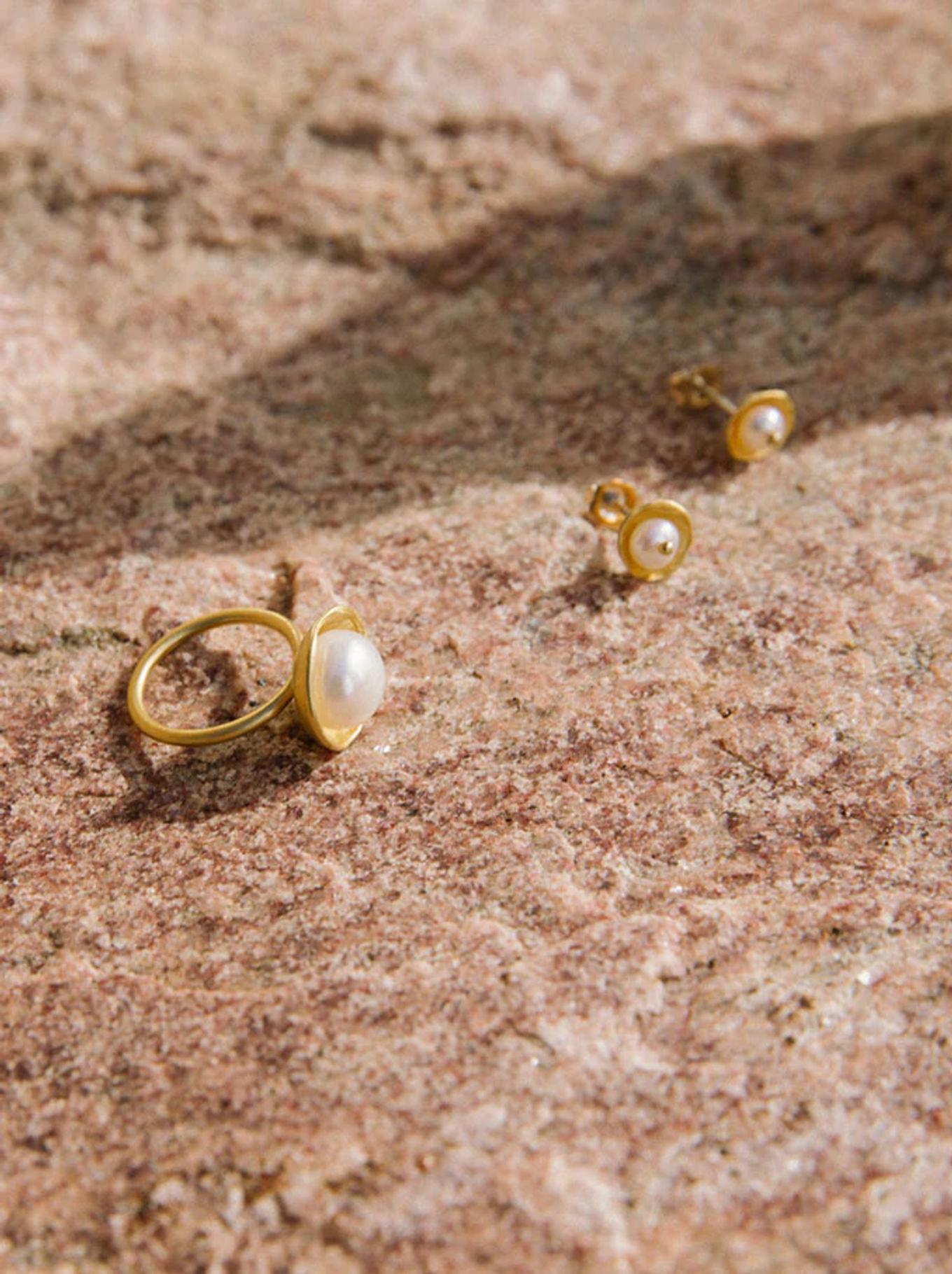 Gold pearl ring and pearl stud earrings on textured stone in natural light. Simple designs showcase jeweller's skill with organic materials.