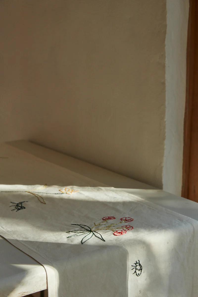 Sunlit beige tablecloth with flower and insect embroidery draped over table edge. Light wall background creates serene breakfast atmosphere.