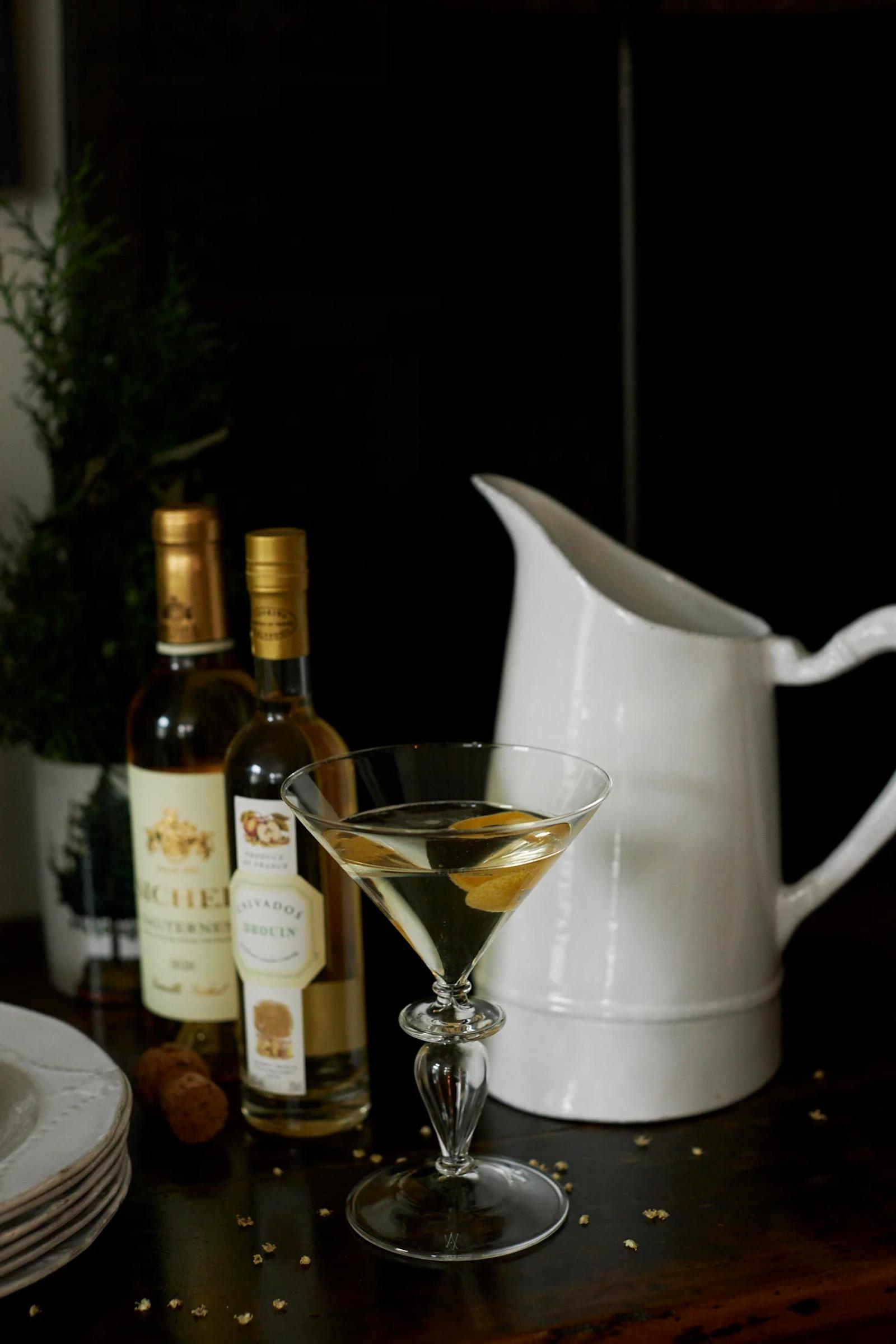 martini glass filled with a tinted cocktail garnished with a lemon twist sits on a wooden table. Behind it, there are two bottles of alcohol, an elegant white ceramic pitcher, and some white plates stacked to the left. Foliage in the background adds a touch of Marseille charm to the scene.