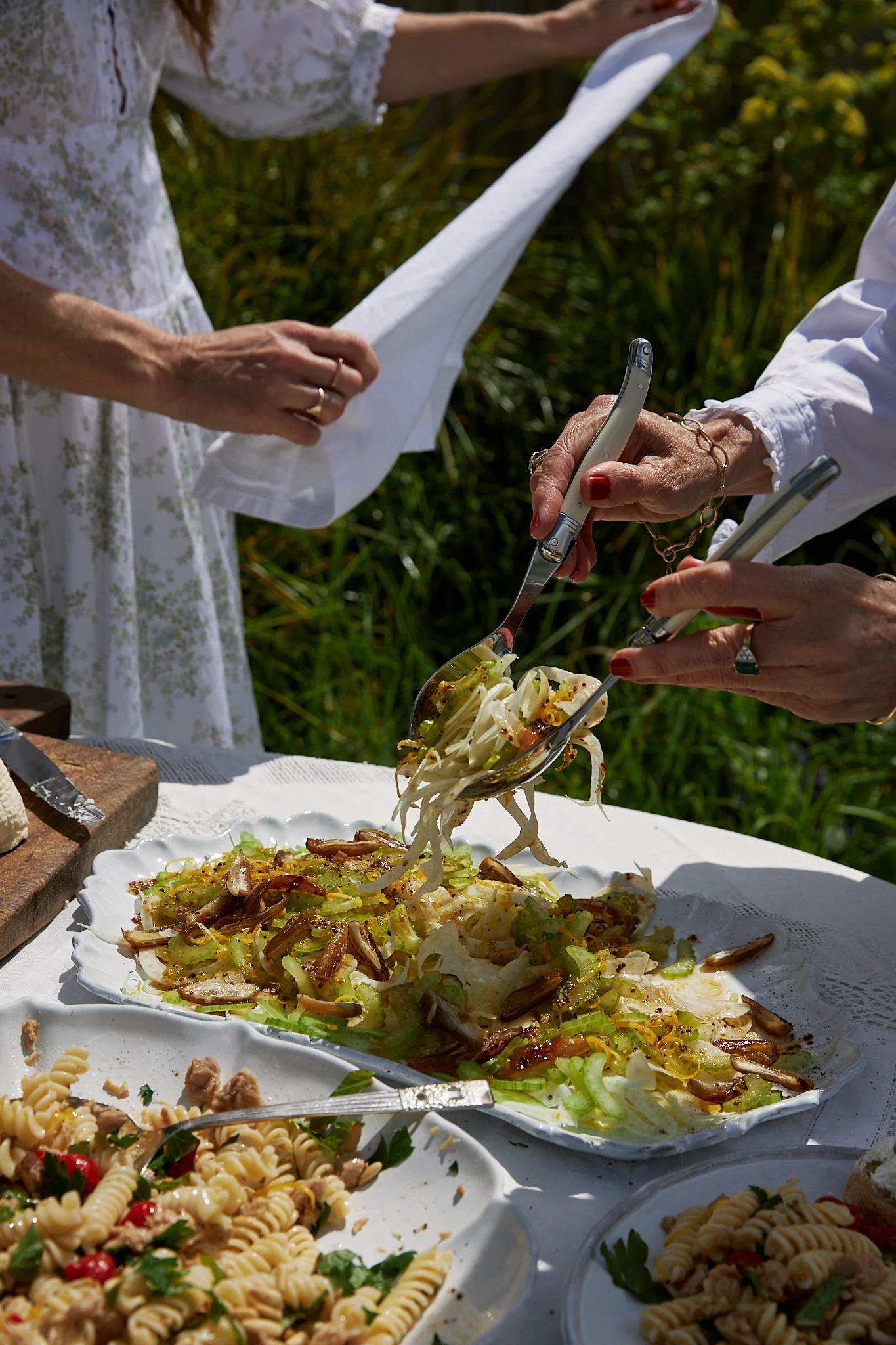 two women stand around the table in the garden, one serves salad onto plates, the other opens a fresh napkin