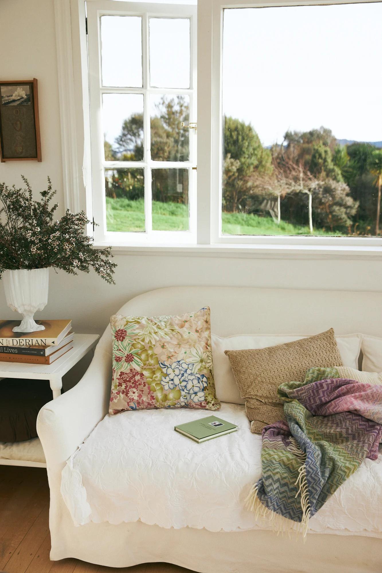 White bench with floral pillows and knitted blanket. Open book on bench. Novels stacked near vase. Windows show garden view.