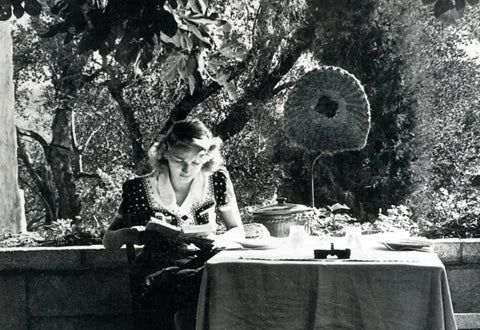 Woman reading outdoors under tree. Sun hat hanging nearby. Table with teapot. Tall trees in background.