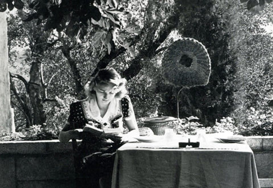 Woman reading outdoors under tree. Sun hat hanging nearby. Table with teapot. Tall trees in background.