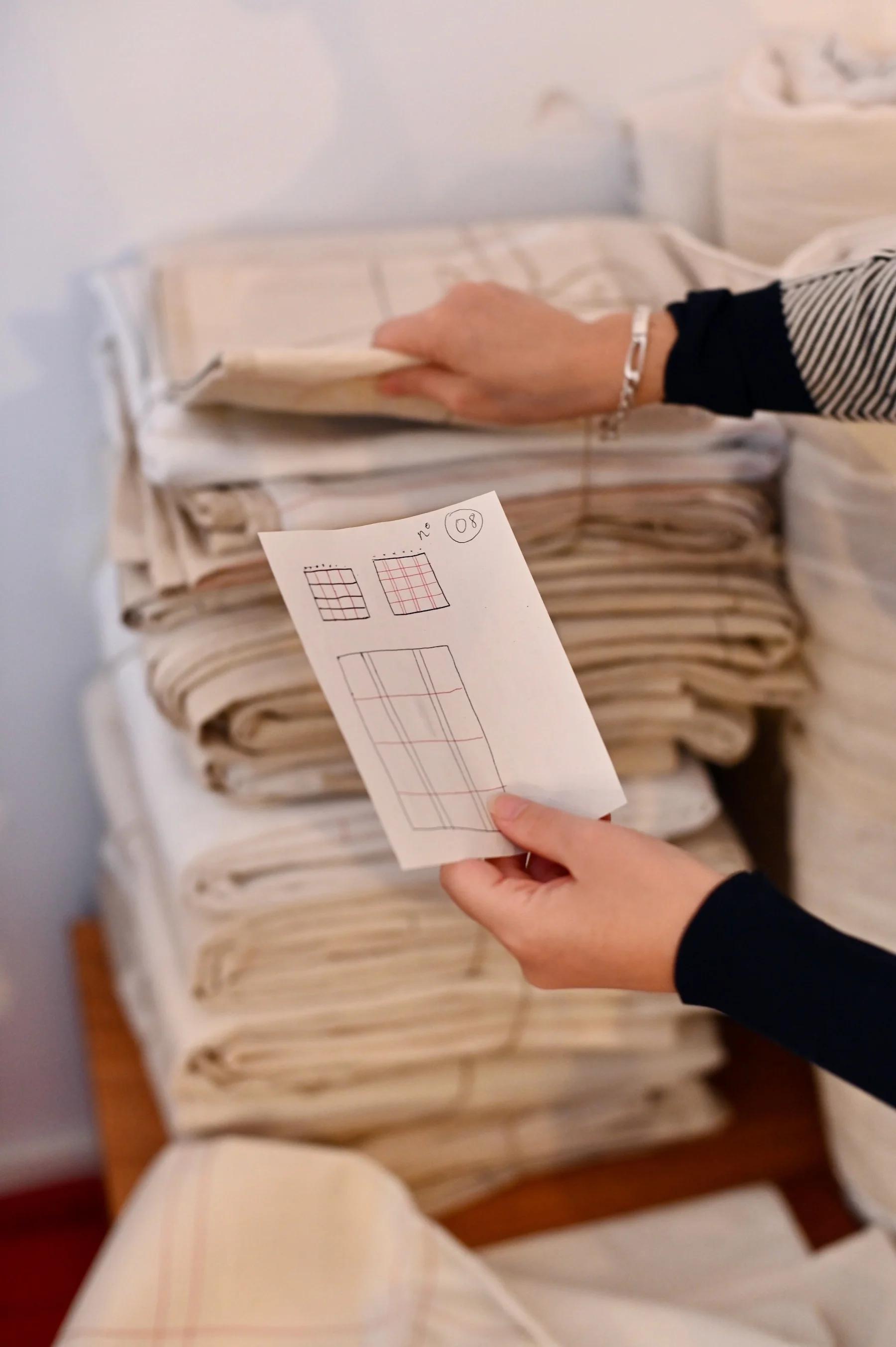 Person in striped shirt holds paper with grid patterns. Folded white and beige fabrics in background.