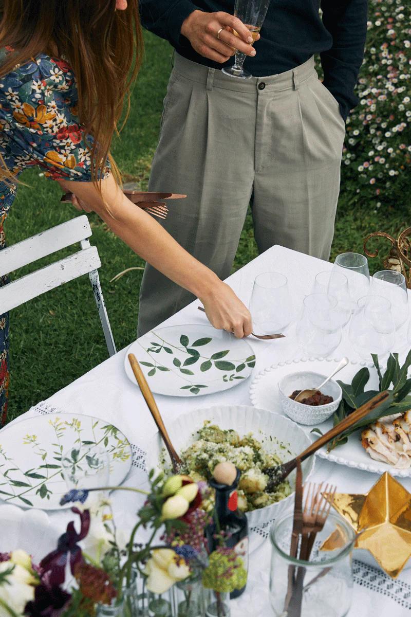 Person in floral dress setting fork on leafy-patterned plates. Another person holds wine glass. Outdoor setting.