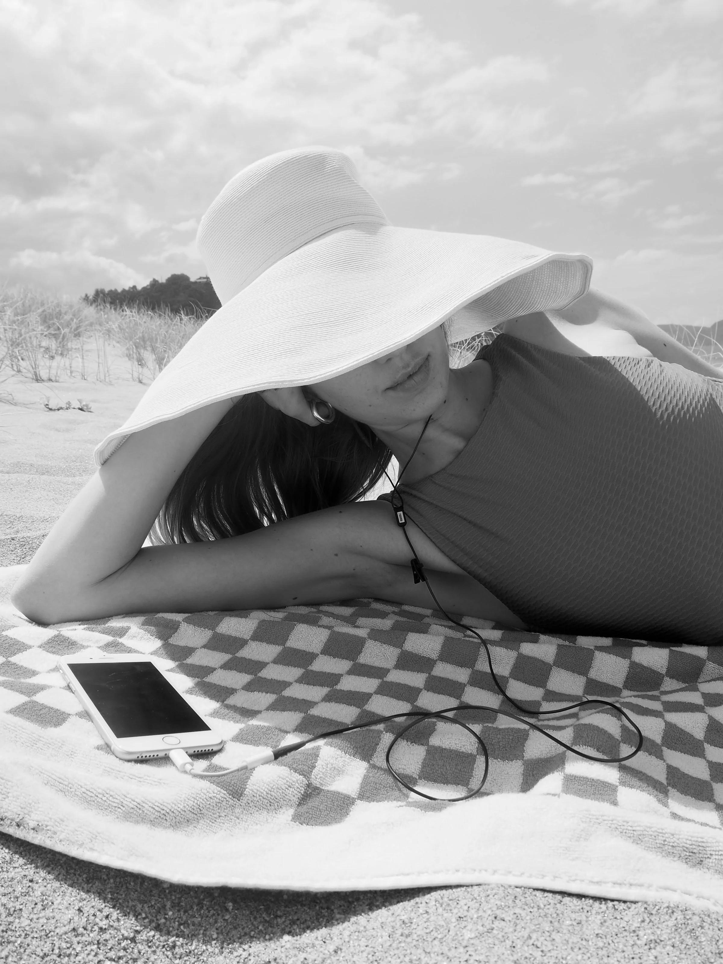 A woman reclines on a checkered towel at the beach, wearing a large sunhat. Earbuds are connected to her smartphone, with dune grasses and a cloudy sky in the background.