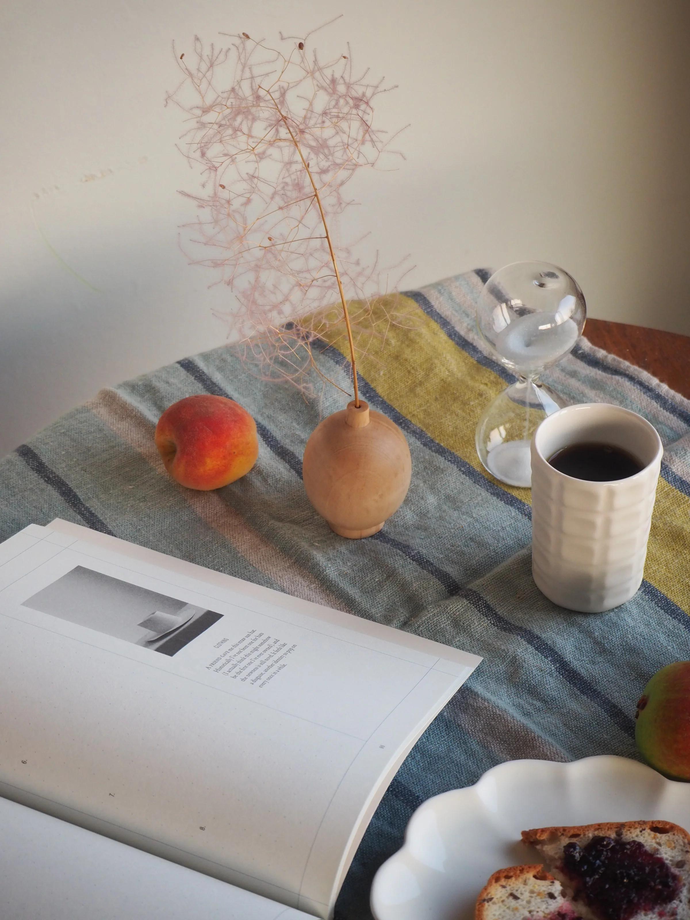 A serene table setting with an open book, peach, dried flowers, toast with jam, coffee cup, and hourglass on a striped cloth at dawn.