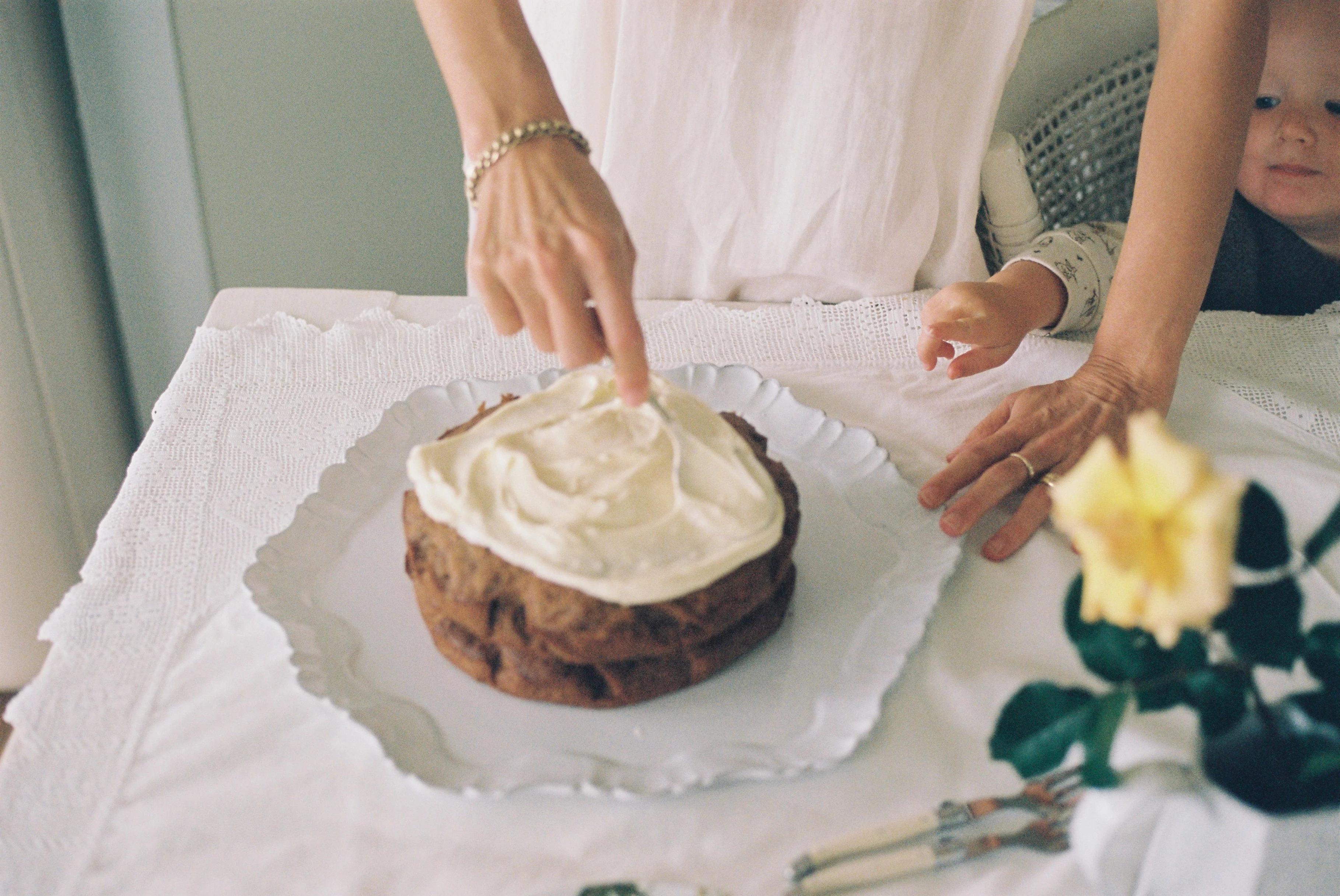 Person icing carrot cake on lace tablecloth. Yellow rose in vase. Child's hand reaching towards cake.