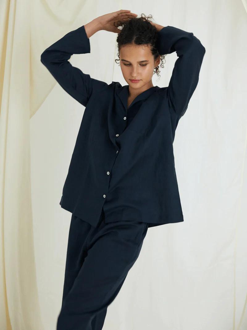 Person in dark blue cotton outfit stands with arms raised. Curly hair, white curtain background. Relaxed, introspective pose.