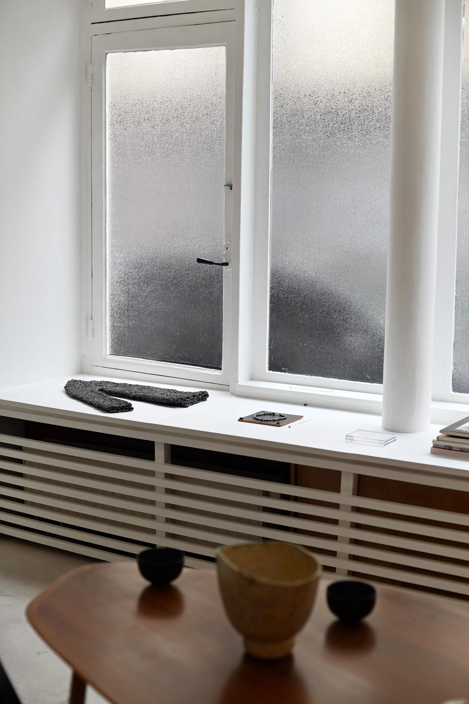 Frosted glass window with white frames. White slatted bench below with grey blanket and magazine. Wooden table with bowls in foreground.