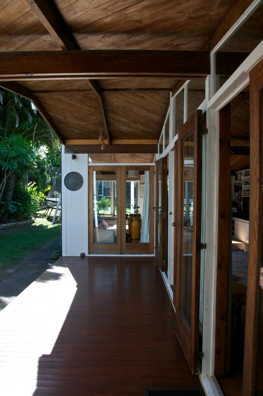 Angled-roof veranda with open glass doors leads to green backyard. Seating and decor visible inside. Light filters through, highlighting vibrant details.