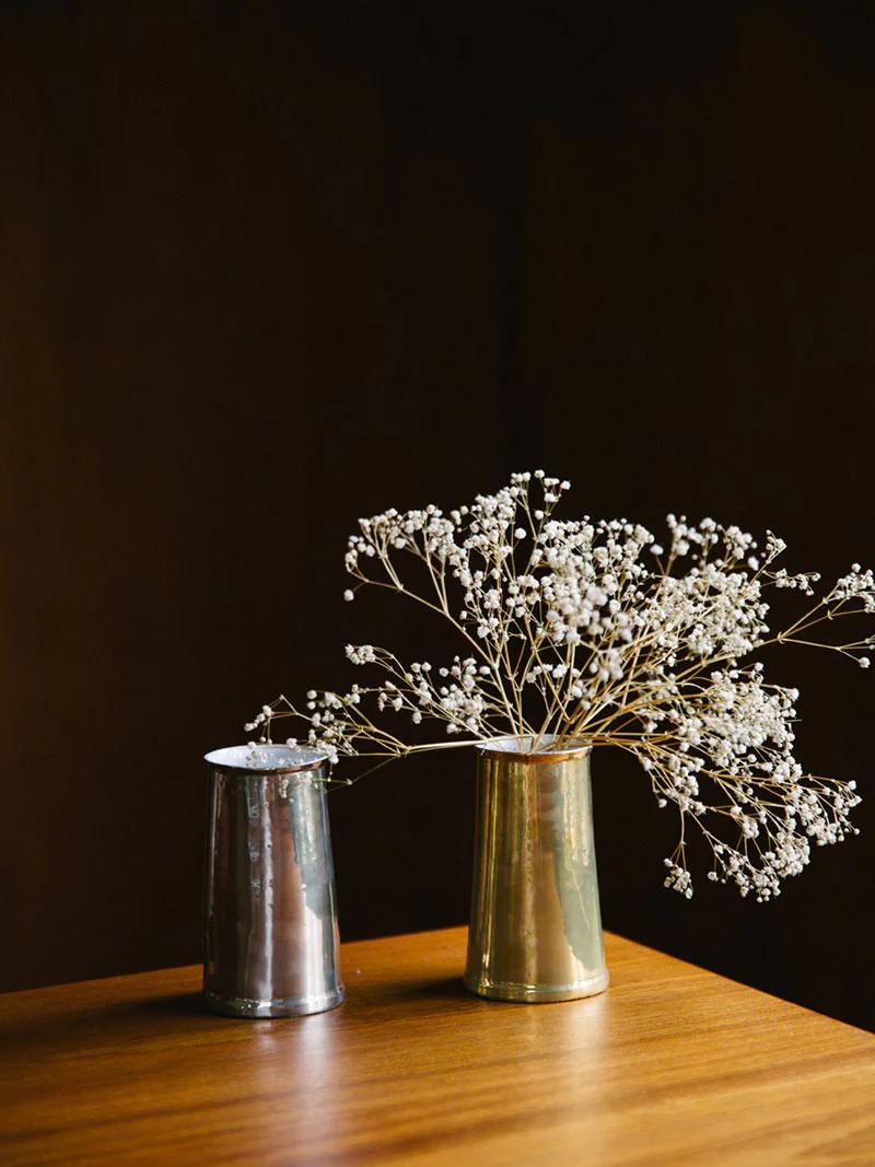 Contrasting metal vases on wooden table: Small silver vase alongside larger gold one holding delicate white baby's breath. Dark background enhances Parisian boutique ambiance.