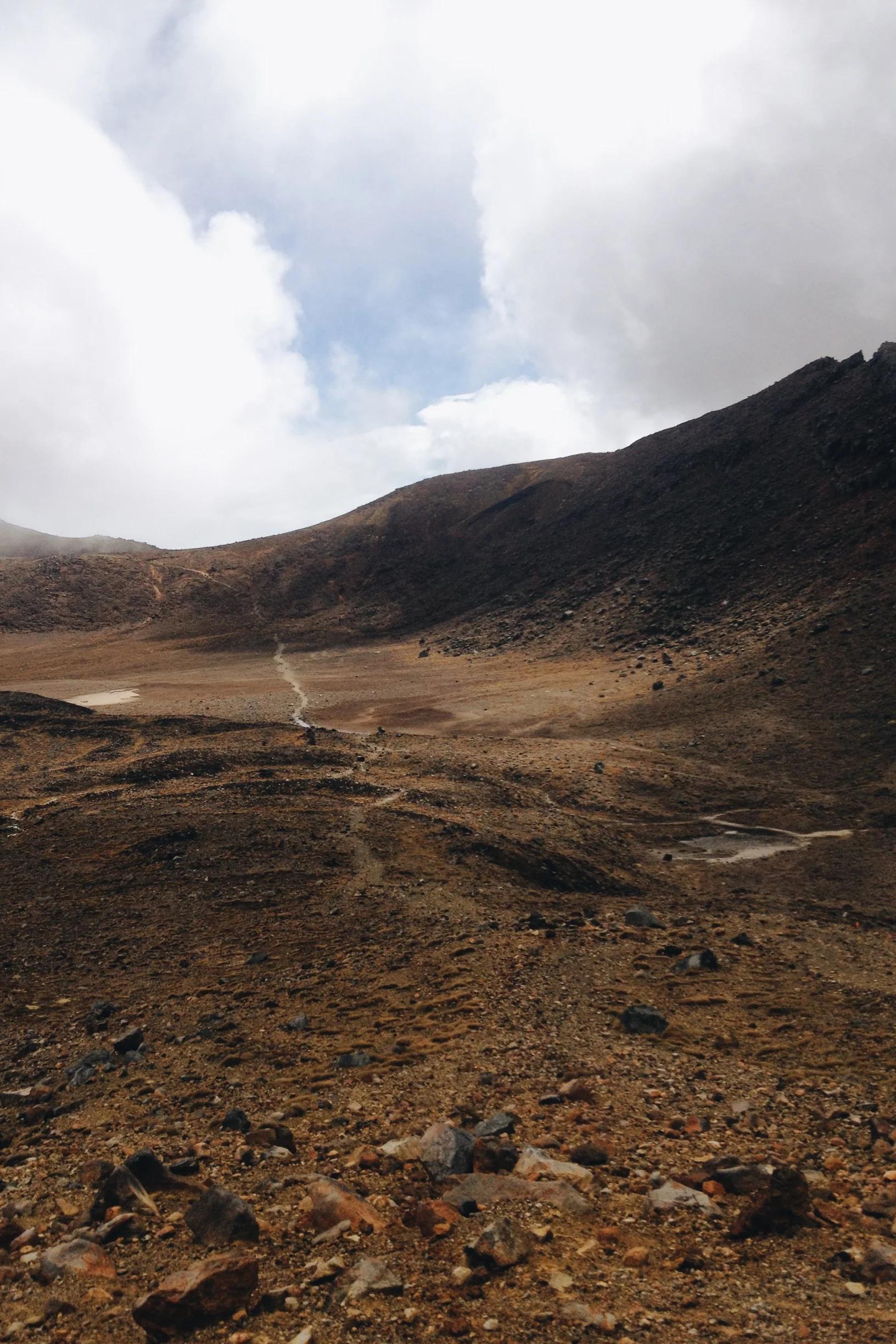 Desolate desert landscape with rocky terrain and sparse vegetation. Distant hills under cloudy sky with blue patch visible. Off-the-beaten-track scene with depth and contrast