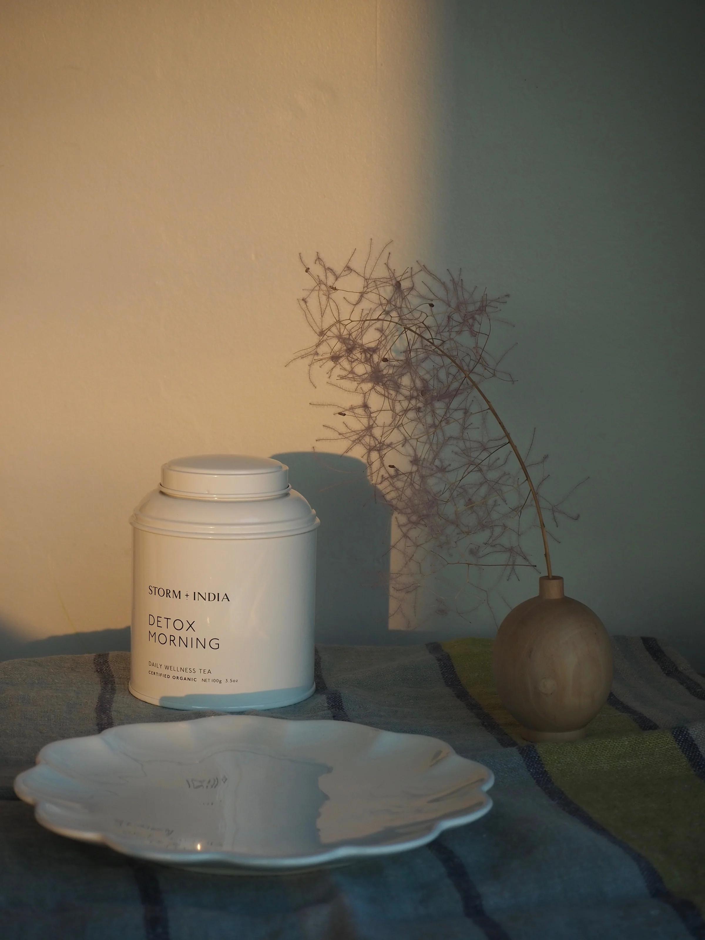 A minimalist setup with a tea canister, wooden vase, and scalloped plate on a striped cloth, illuminated by soft morning light.