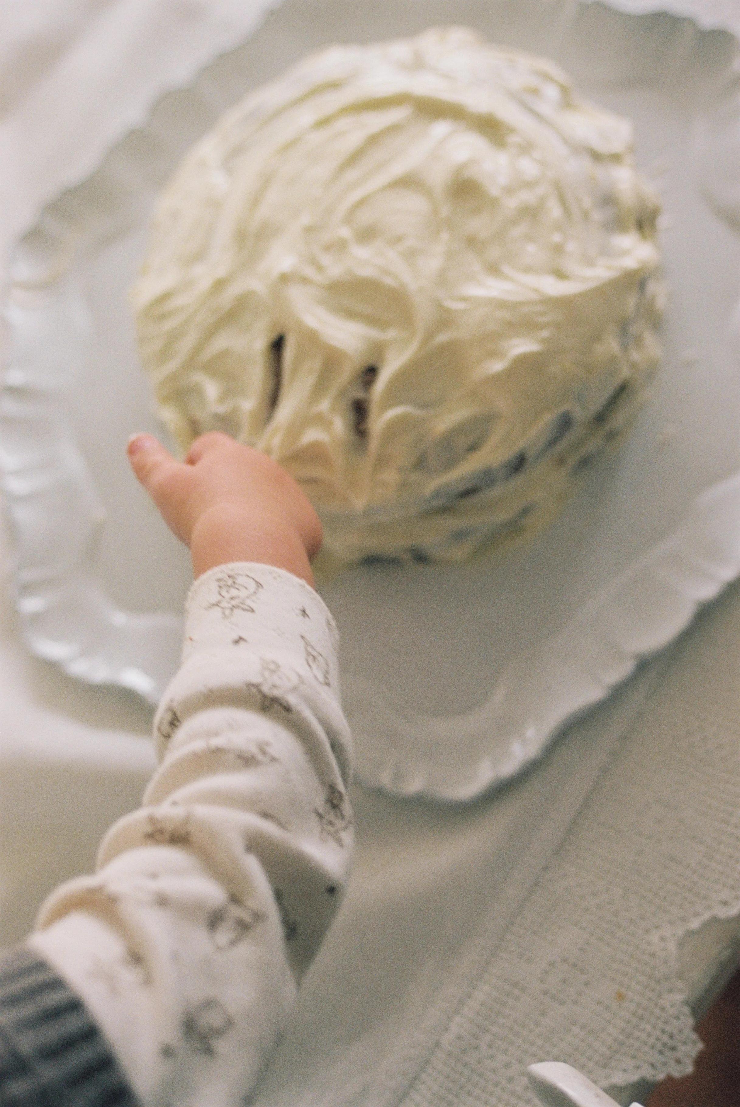 Child's hand touching frosted carrot cake on decorative plate. Arm and patterned sleeve visible.