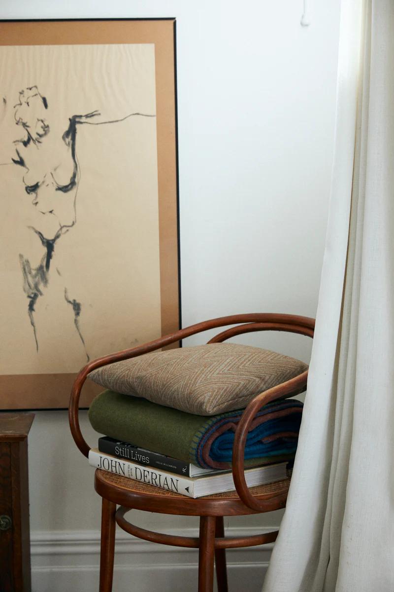 Wooden chair holding folded blankets in earthy tones. Three books on top, "John Derian" visible. Framed sketch leans against wall.