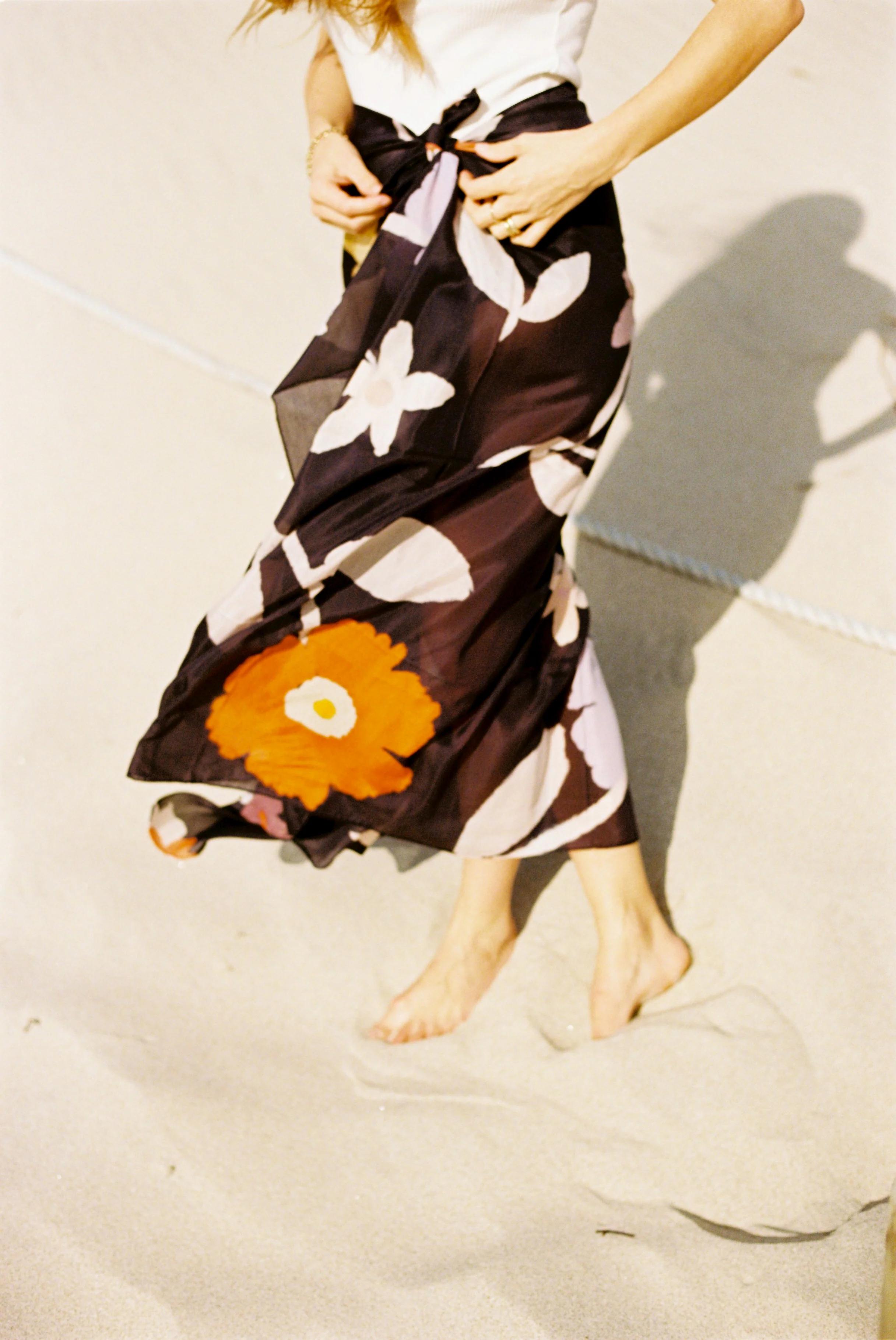 Person walking barefoot on beach. Flowing black skirt with large floral patterns. White sleeveless top. Face out of frame.