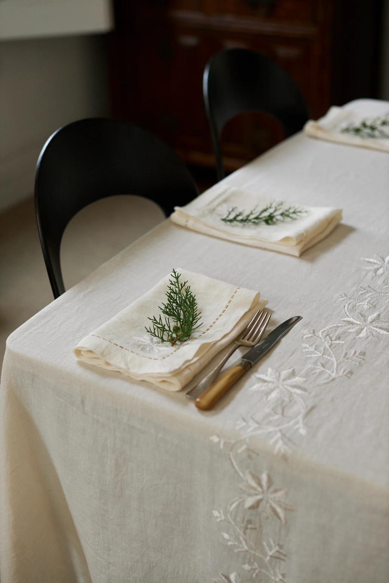 A dining table with three place settings on an embroidered cloth. White napkins with greenery and cutlery are set, with black chairs partially visible