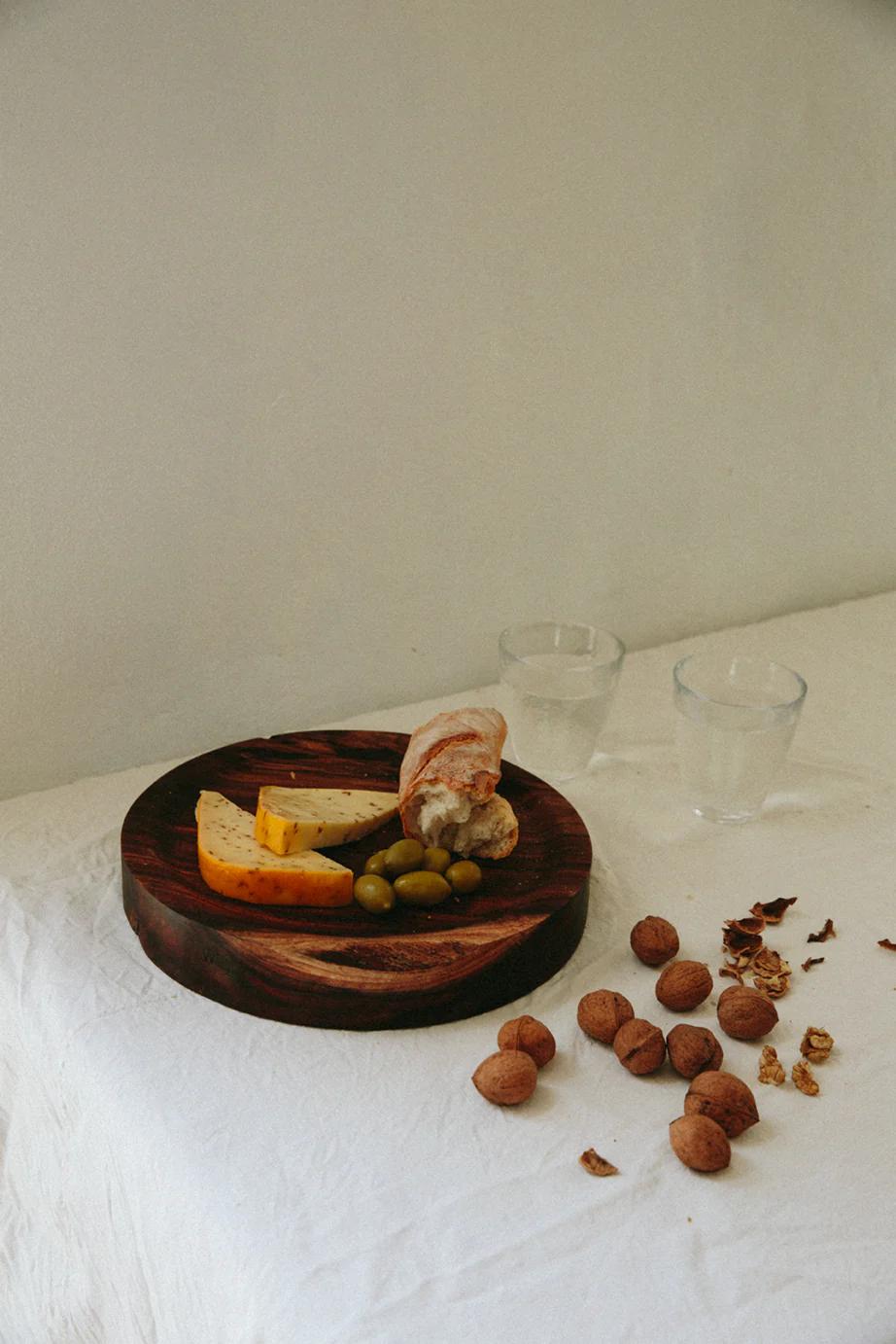 Wooden platter with three cheeses, crusty bread, and green olives on white tablecloth. Two empty glasses and scattered walnuts nearby. Platter crafted from local NZ wood using traditional techniques.