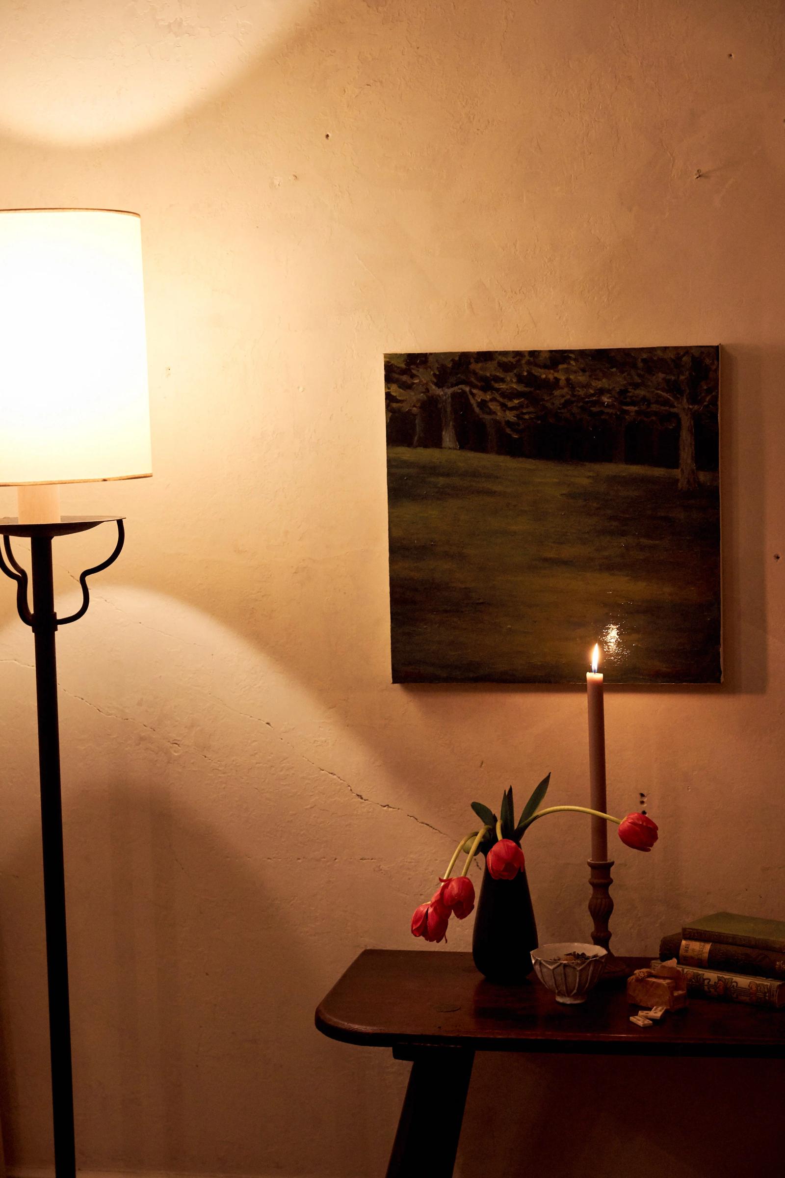 A standing lamp in the corner of a room glows across the walls. A small wooden table with flowers and a lit candle, and a hanging picture are on the right.