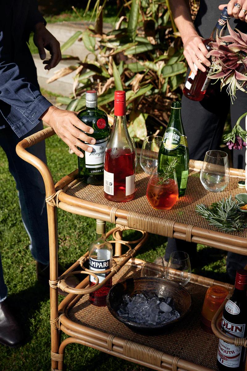 Outdoor wicker bar cart with various bottles and pink drinks. Two people reaching for bottles amid garden party setting.