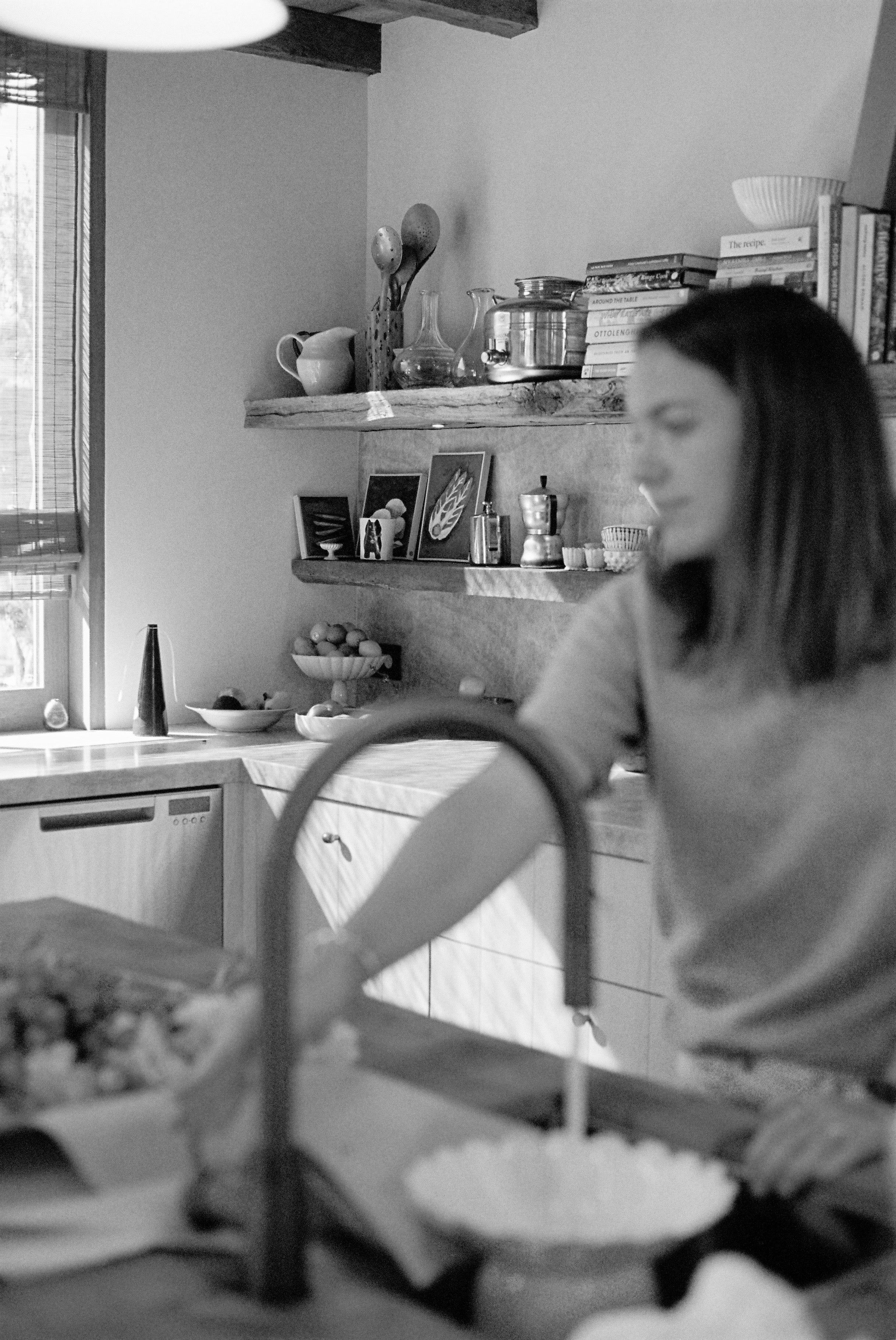 Black and white photo of blurred woman in modern kitchen. Shelves with fruit bowl, books, and jars. Large window lets in natural light.