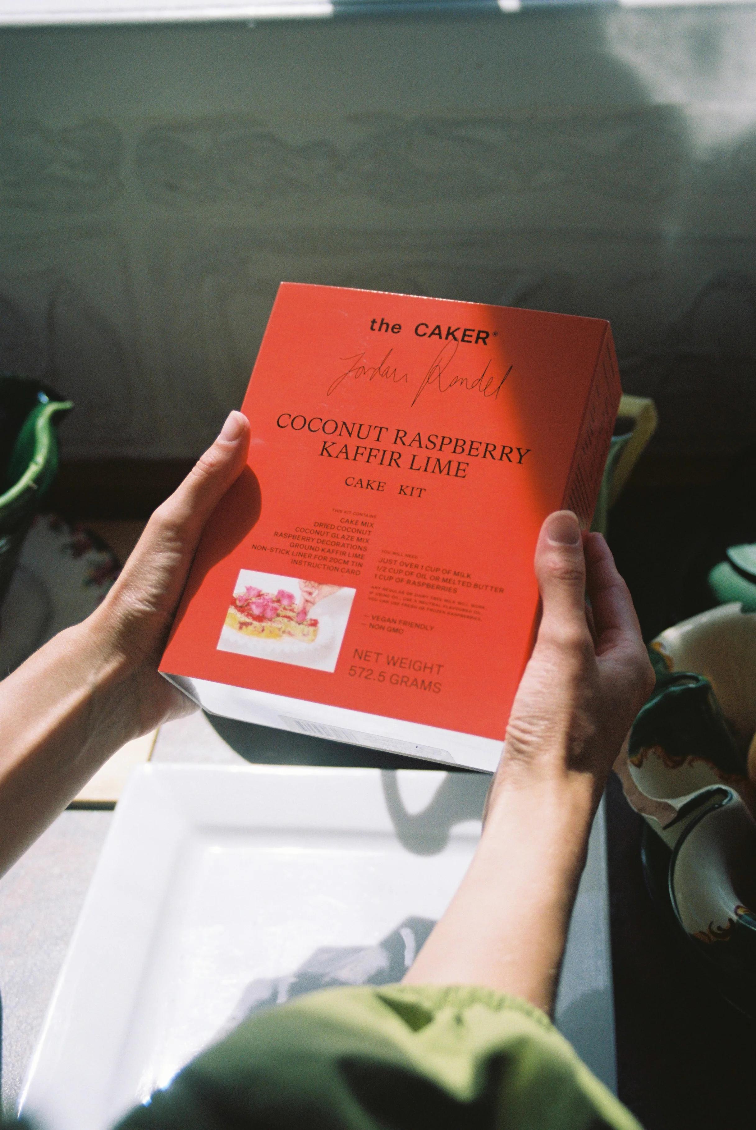 Person holding "the CAKER" Coconut Raspberry Kaffir Lime cake kit box. Bright kitchen setting with kitchenware visible.