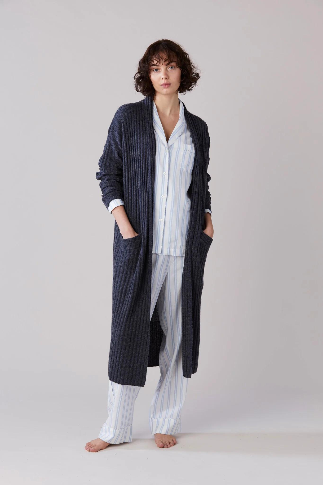 Person in long dark cardigan over striped pajamas. Barefoot on plain background, hands in pockets, short curly hair.