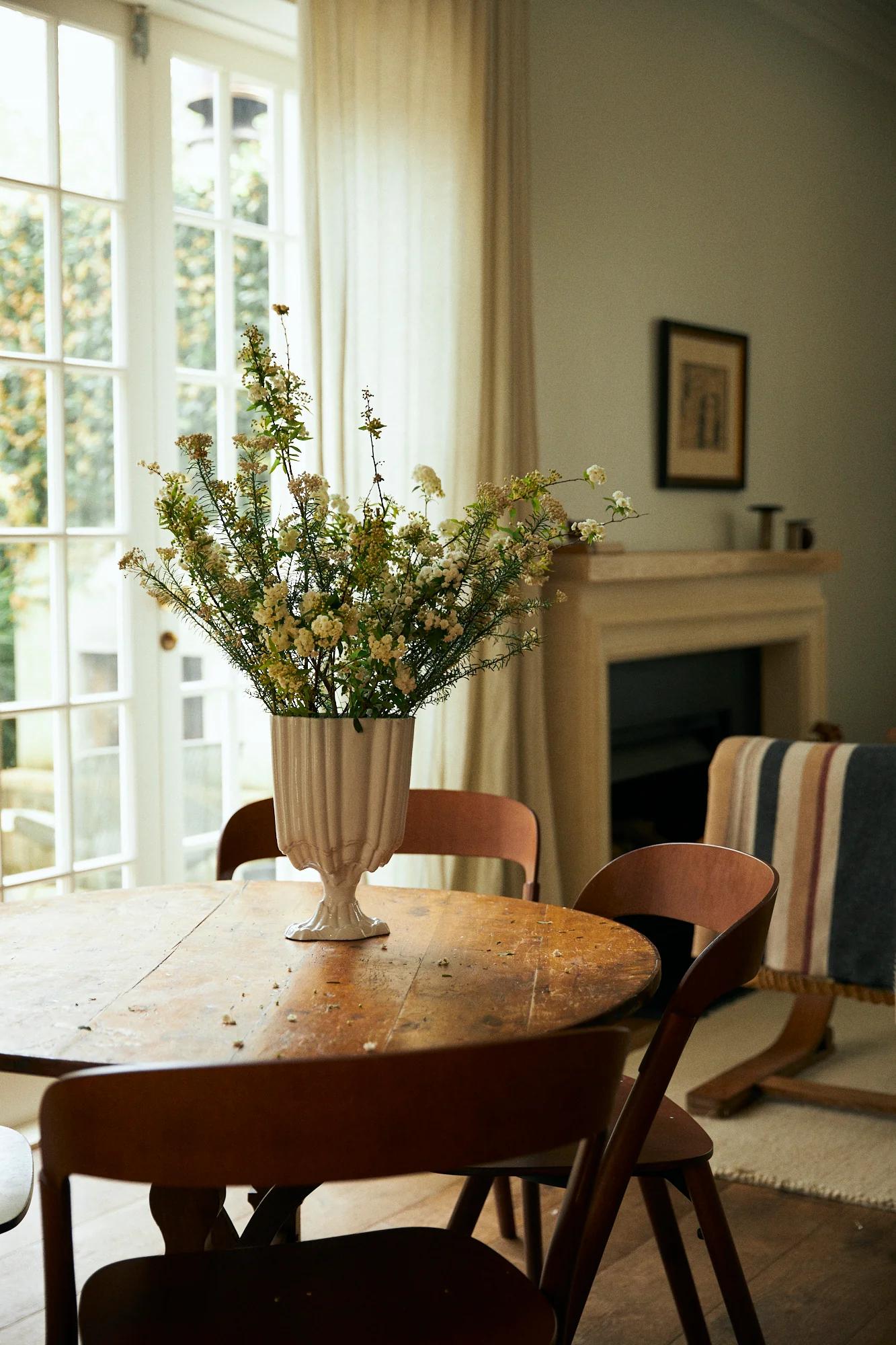 Wooden table with flower vase, surrounded by chairs. Fireplace with artisanal pieces, striped blanket, and large window in background.