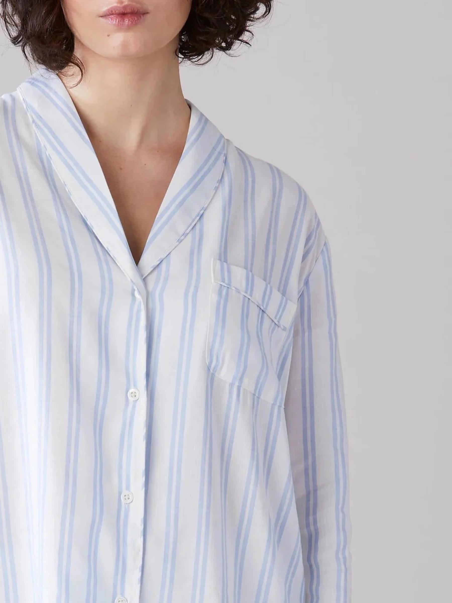 Short-haired person in white shirt with blue stripes. V-neck, buttoned front, chest pocket. Plain light background.